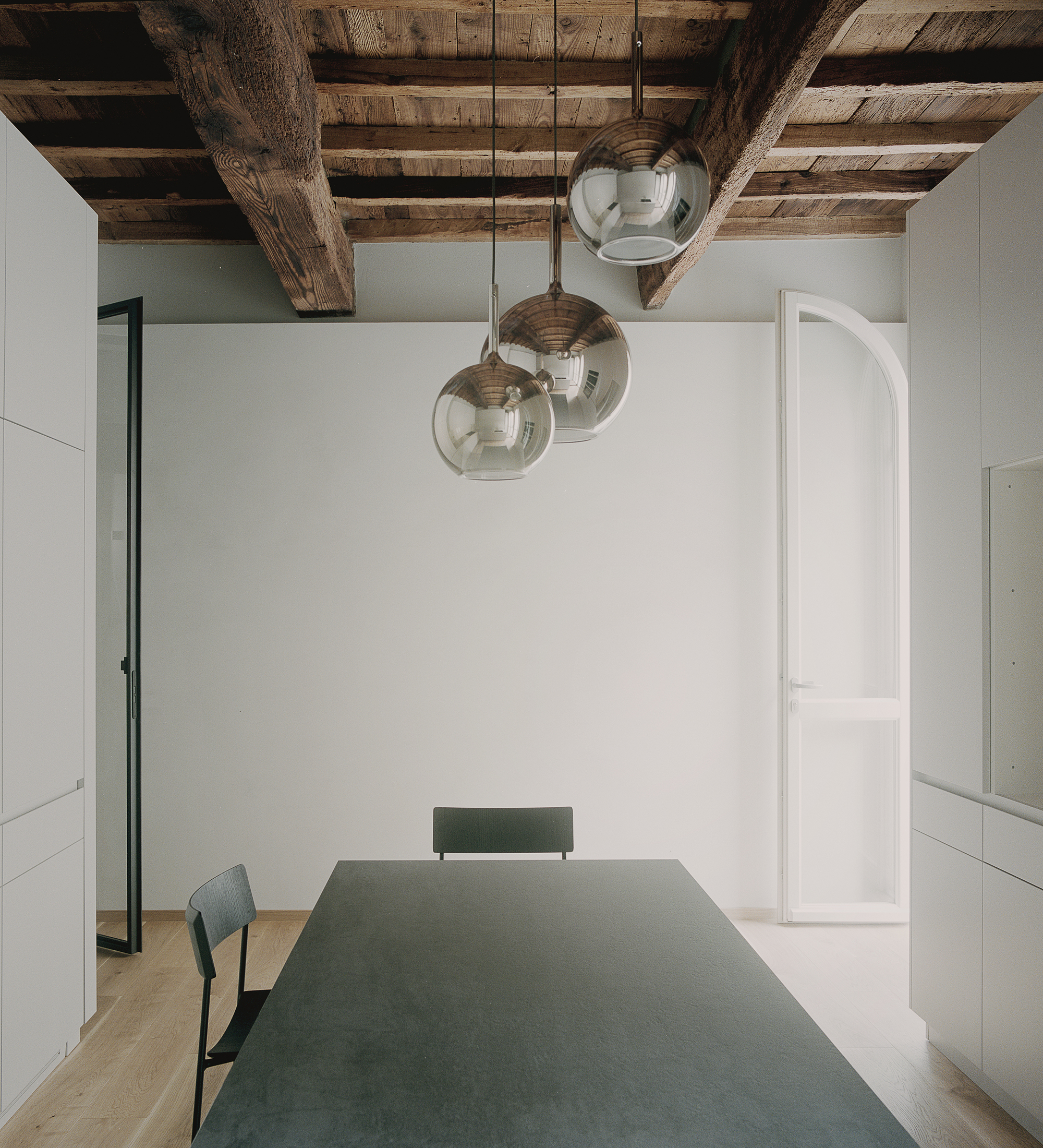 Dining area with green table under three glass pendant lights and exposed timber ceiling beams
