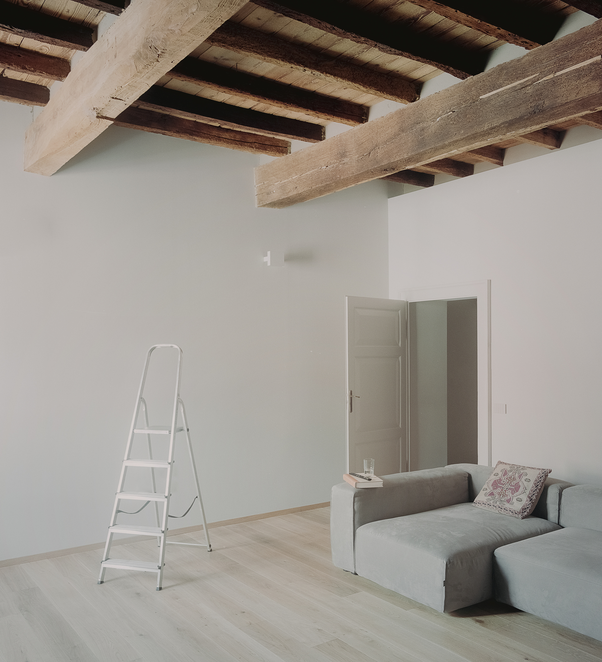 Living room with exposed beam ceiling, open doorway and a stepladder against white walls