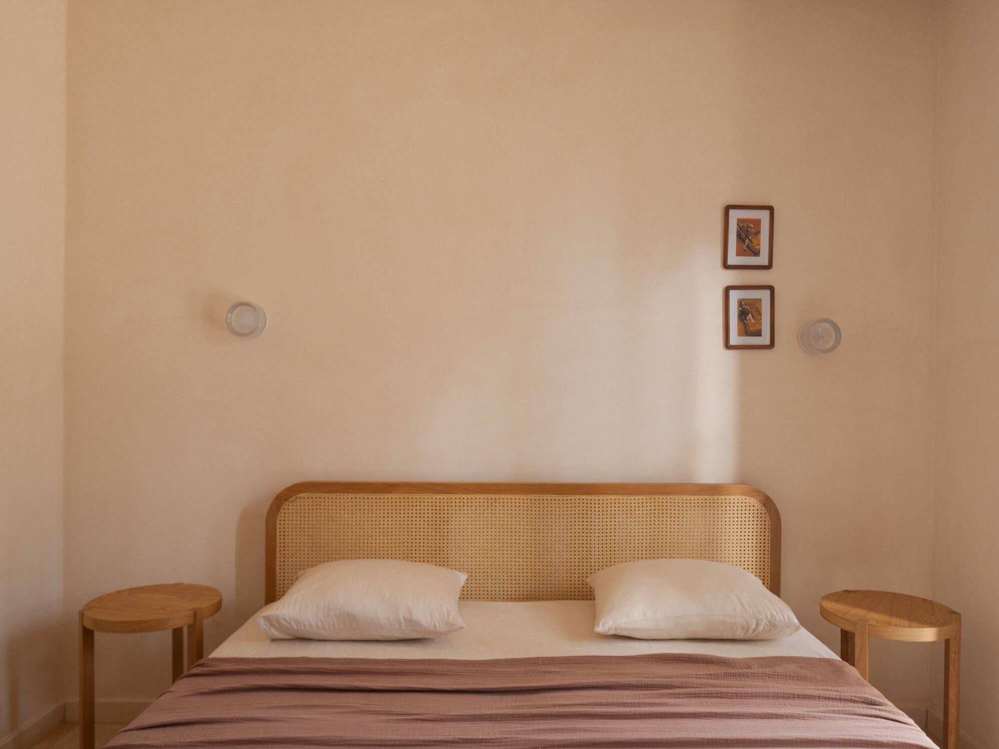Bedroom wall with cane headboard and two framed artworks above cylindrical timber side tables