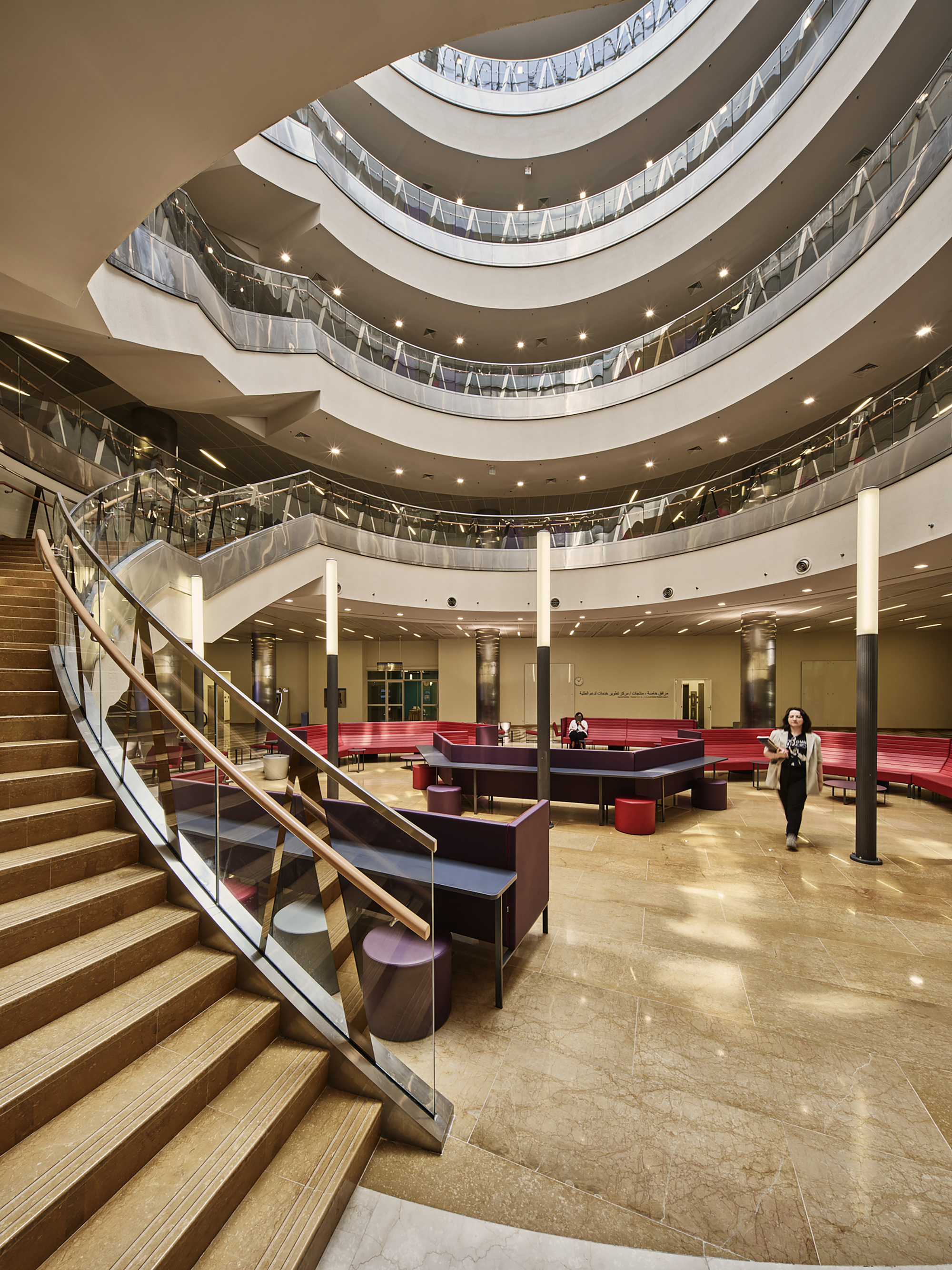 Multi-story atrium with curved balconies, timber and glass railings, and a person crossing the ground floor