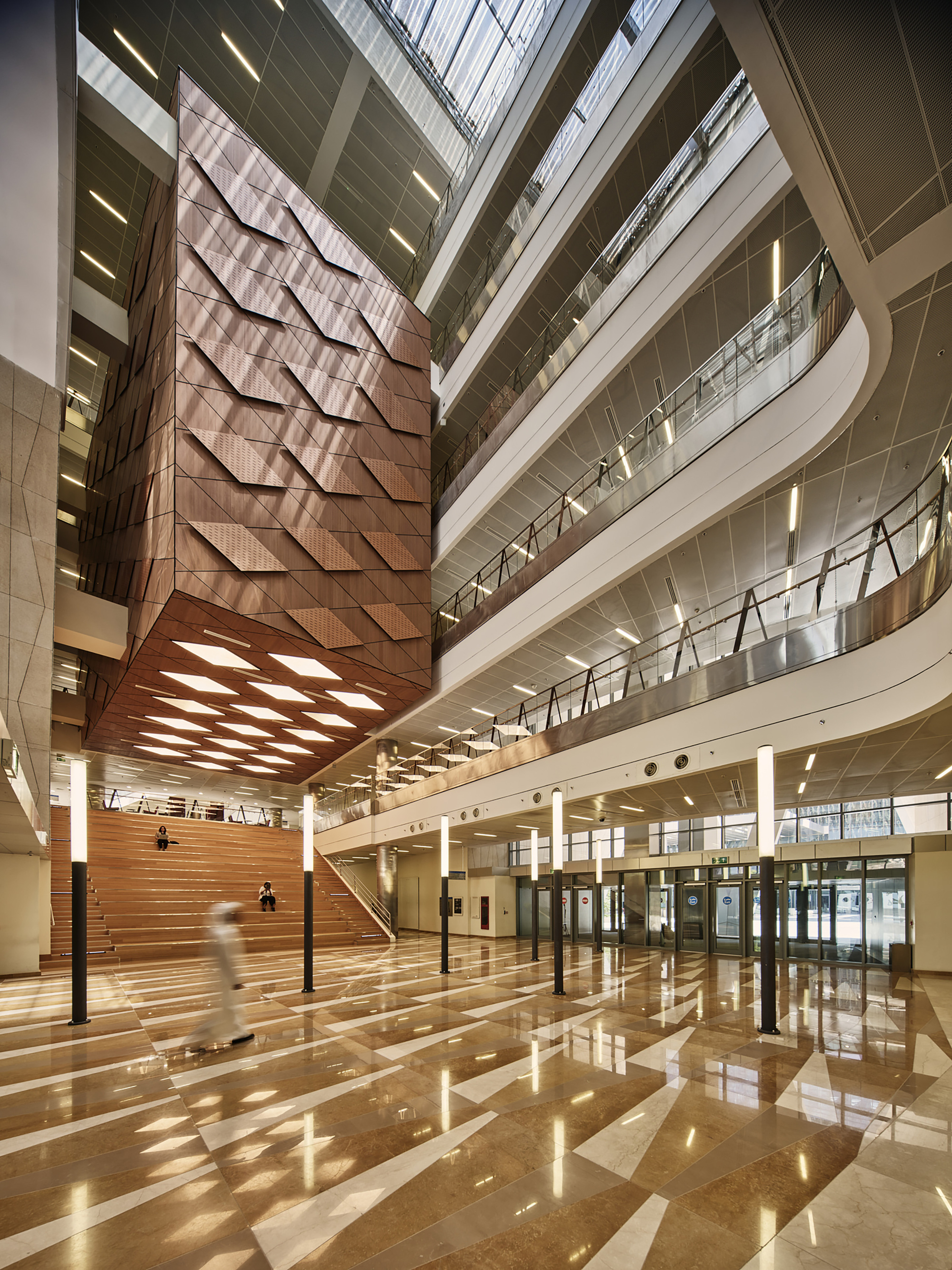 Multi-story atrium with timber-clad seating steps and polished stone floor reflecting skylight patterns