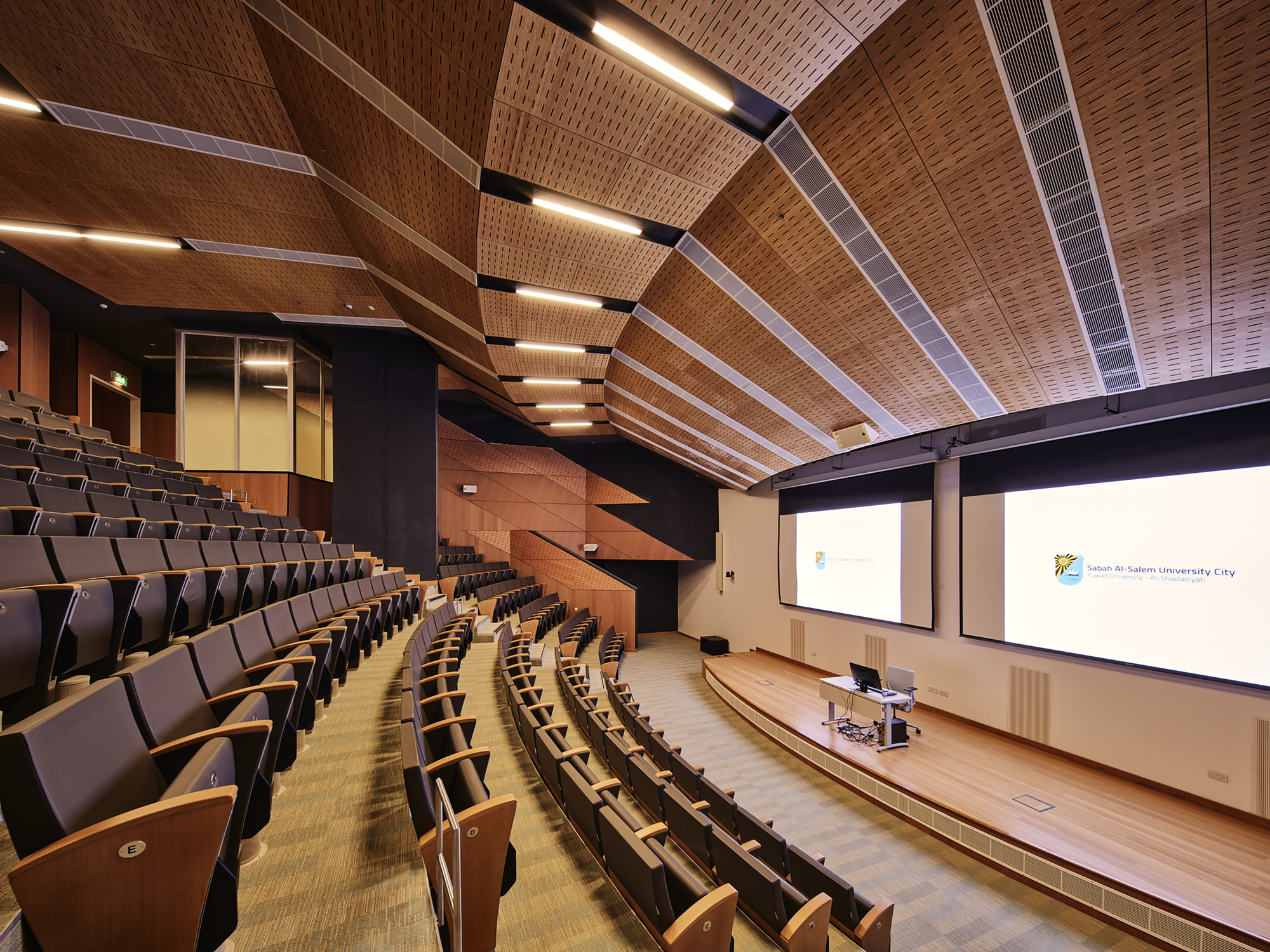 Auditorium interior with tiered seating and woven timber ceiling panels with integrated linear lighting