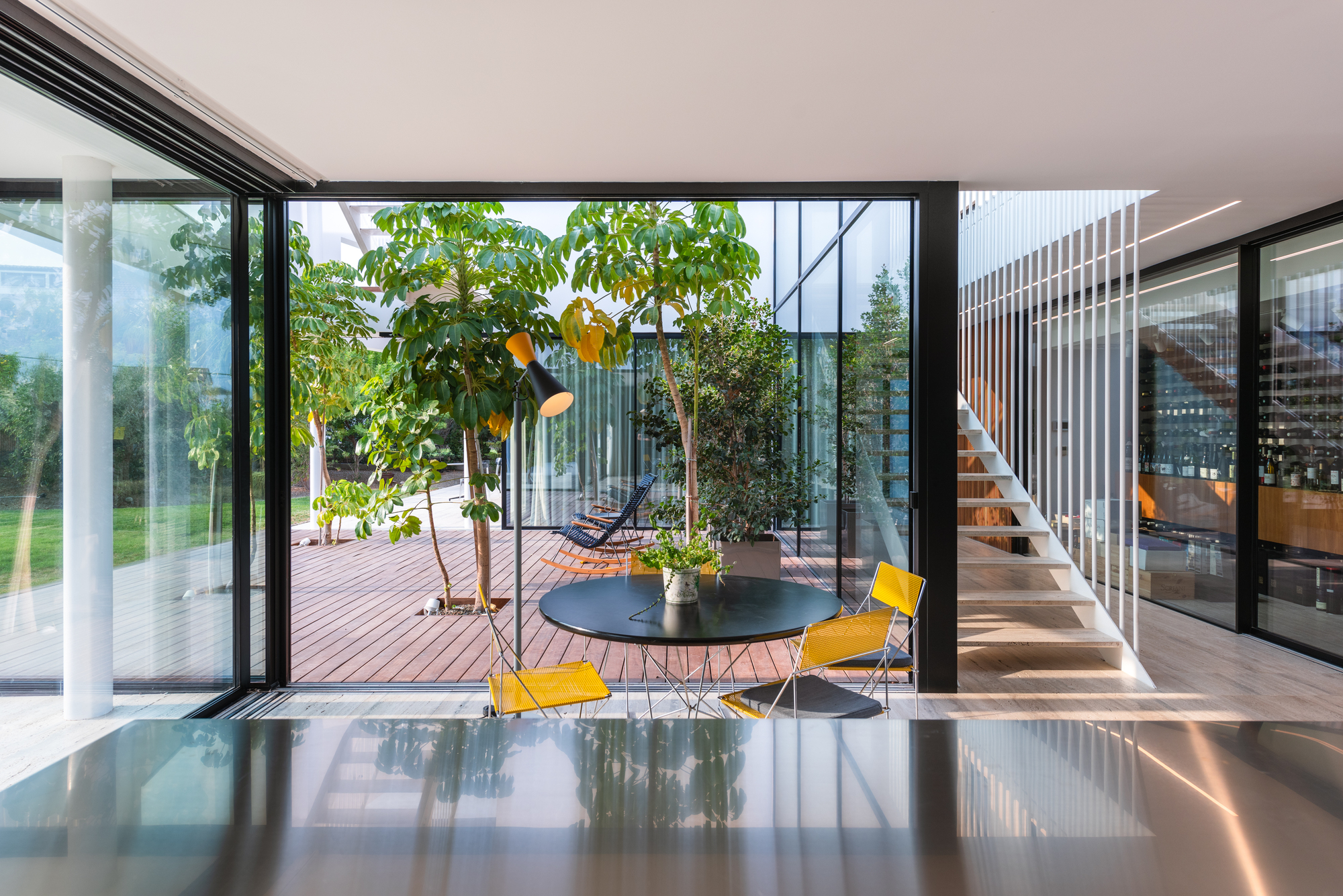 Double-height living space overlooking glazed courtyard with potted trees and timber decking at dusk