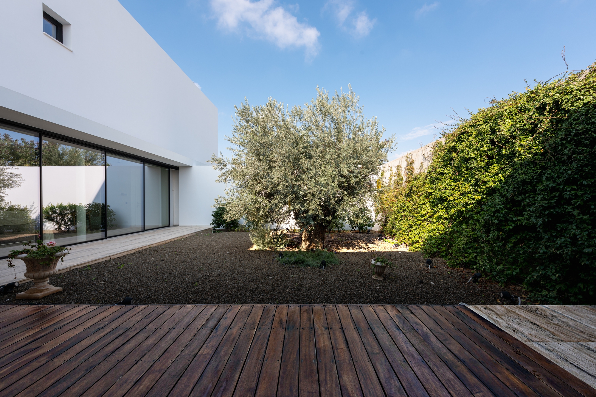 Gravel courtyard with timber deck and olive tree between white plaster volumes under blue sky