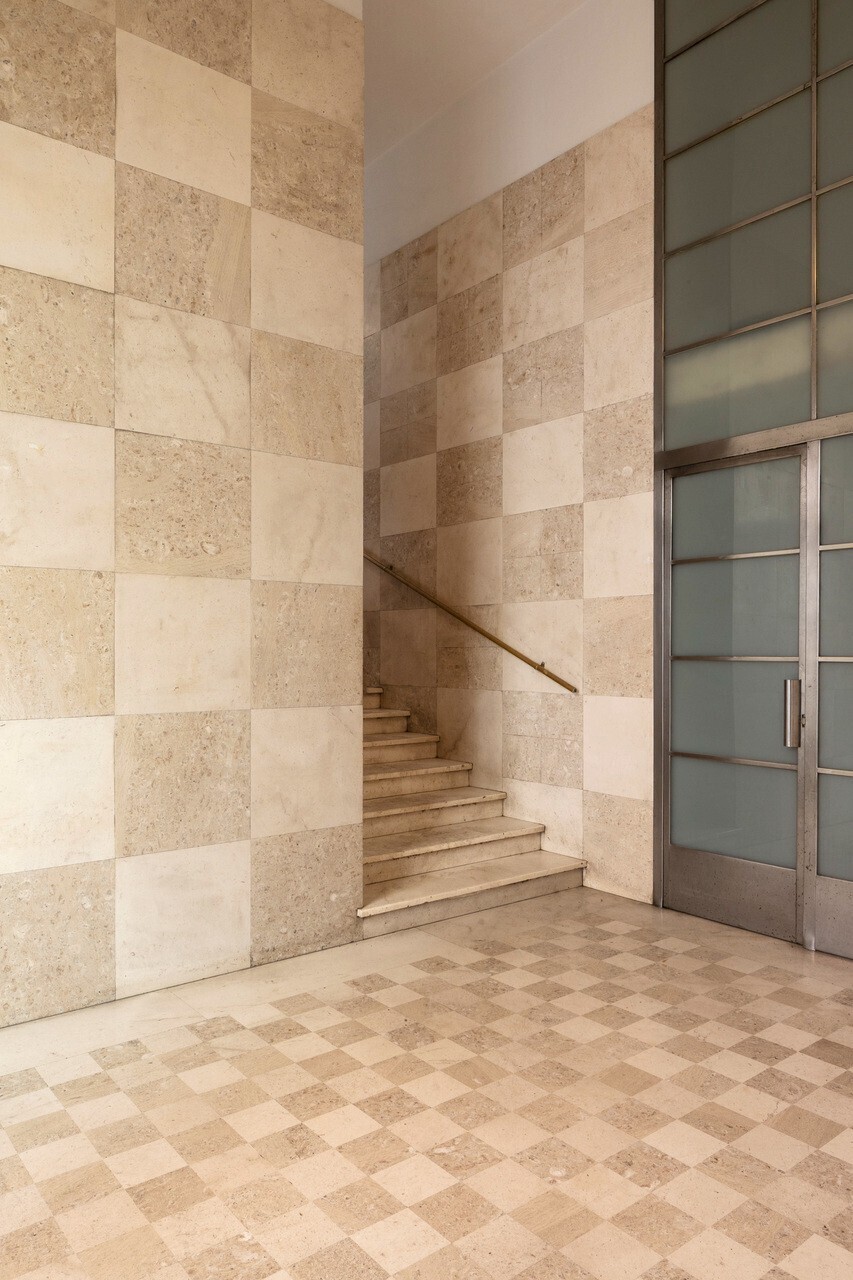 Chequered travertine staircase rising along walls clad in large alternating stone tiles beside a gridded metal door