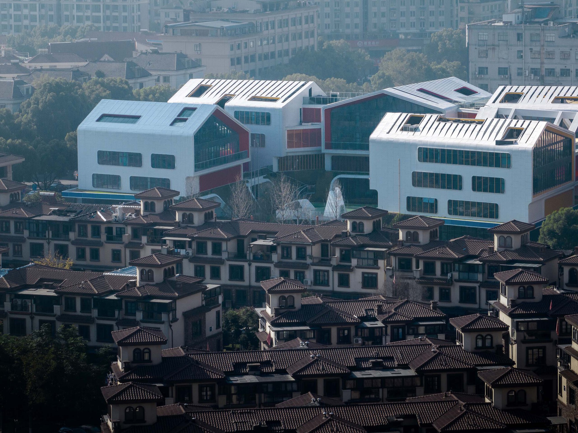 Elevated view of white volumes with louvered roofs rising above a dense neighborhood of tiled-roof houses