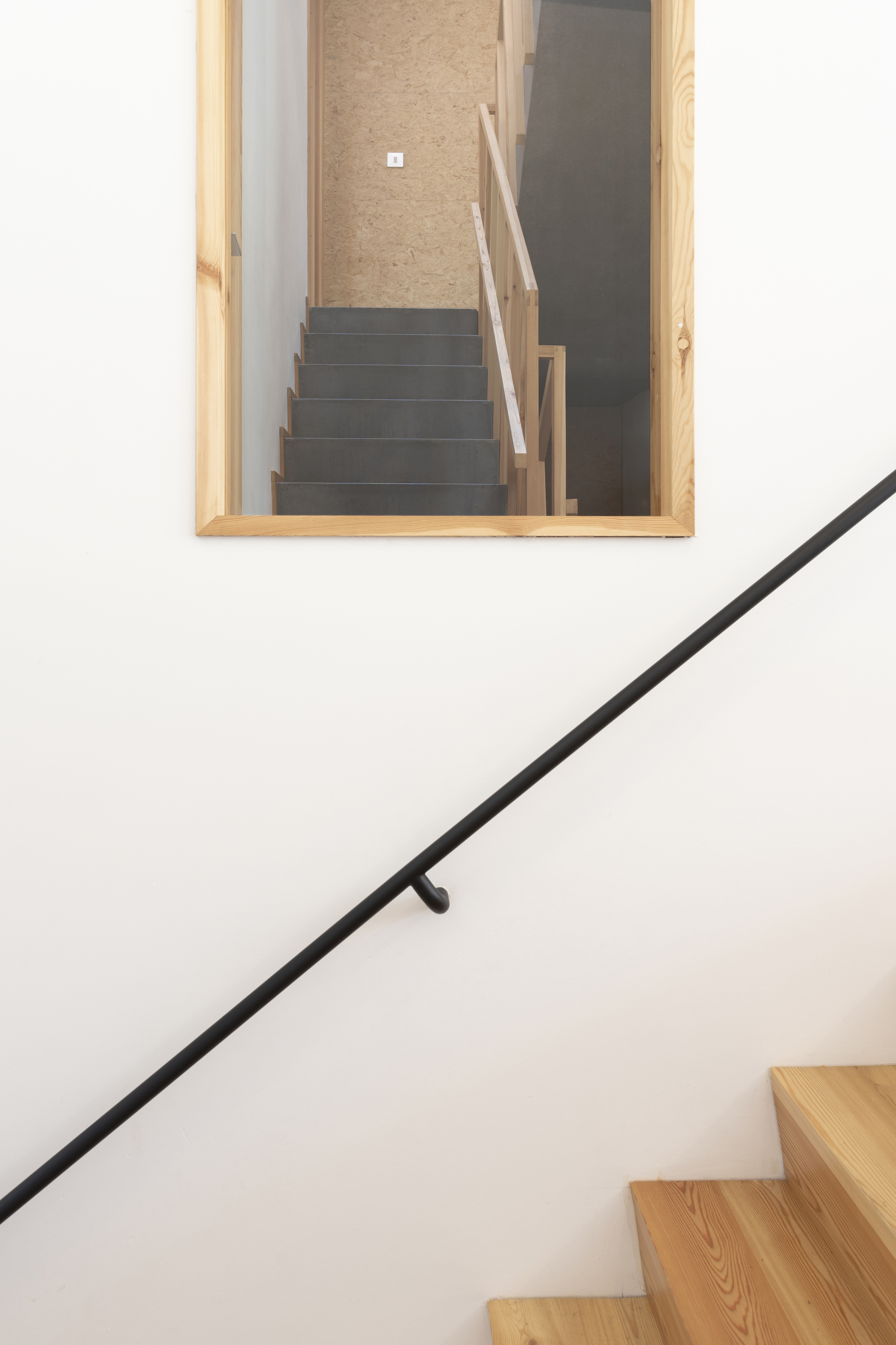 Stairwell with black metal handrail against white wall and timber-framed opening revealing upper level