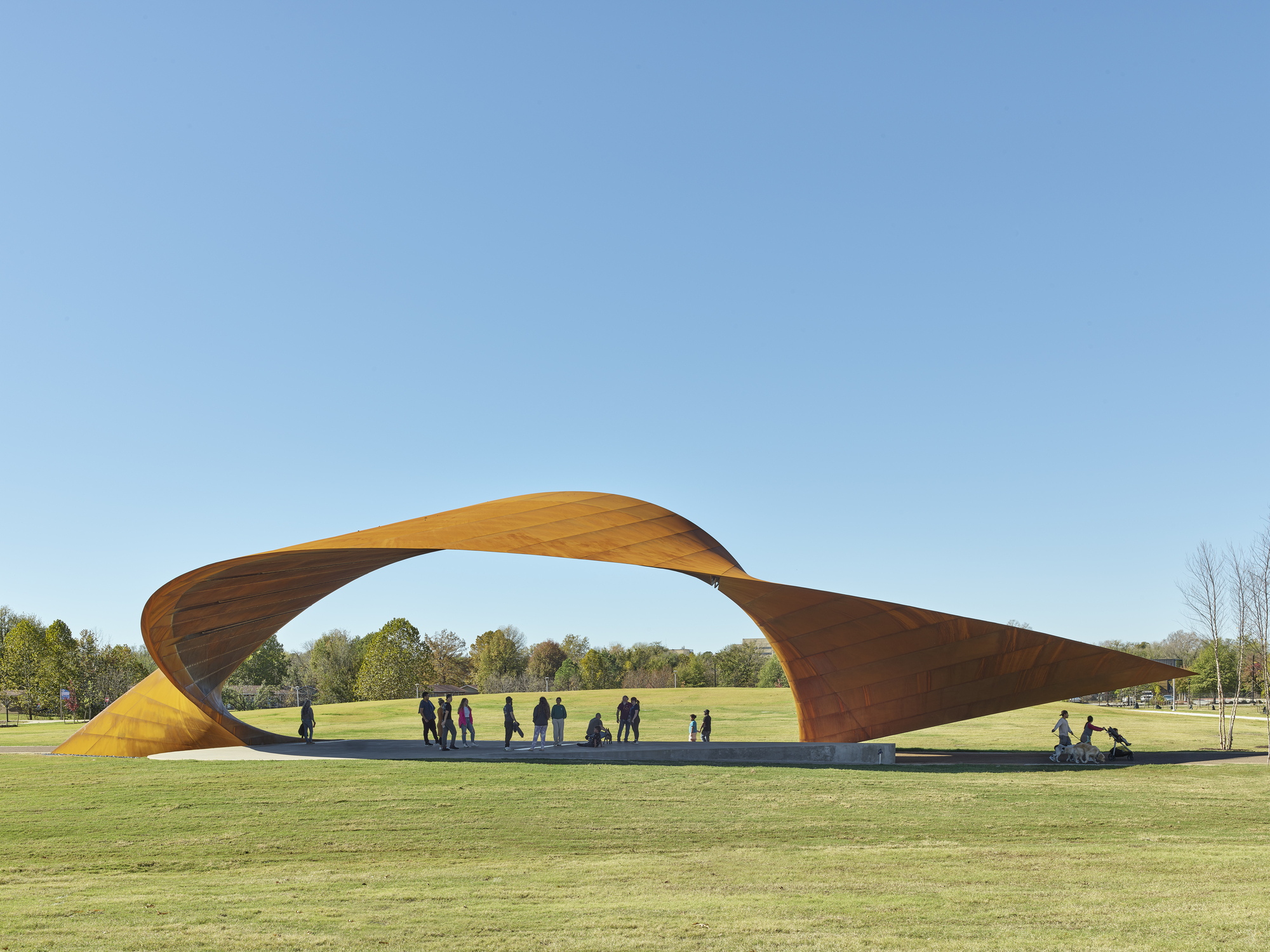 Twisted weathering steel canopy spanning a lawn with visitors gathered beneath on a clear afternoon