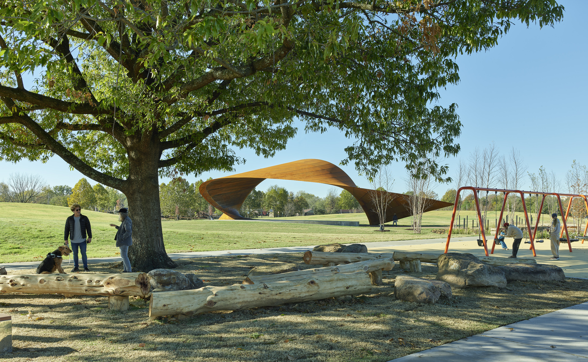 View across natural log play area toward the canopy structure framed by a mature tree