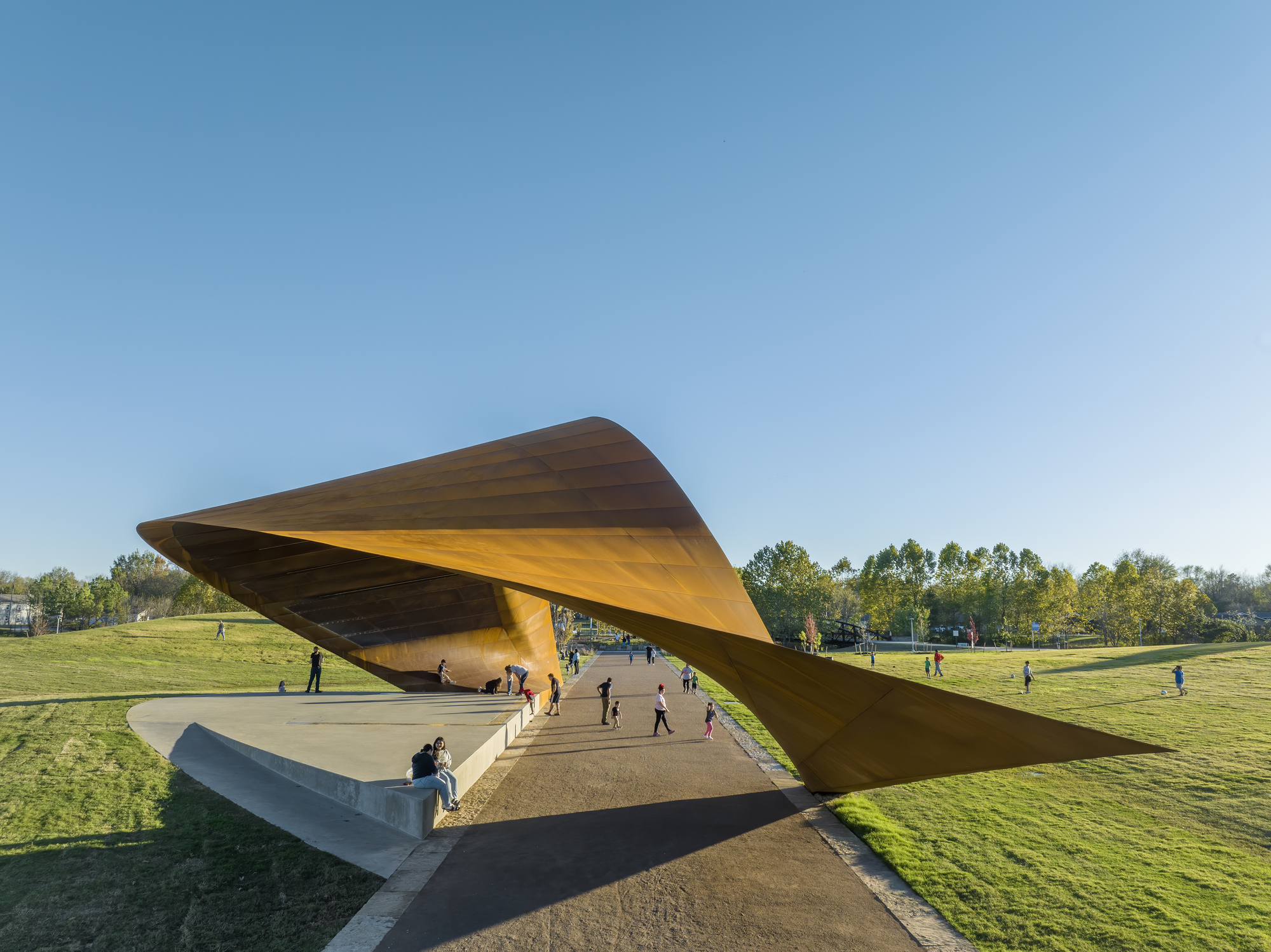 Elevated concrete plaza beneath the lifted steel canopy with families walking in late afternoon light