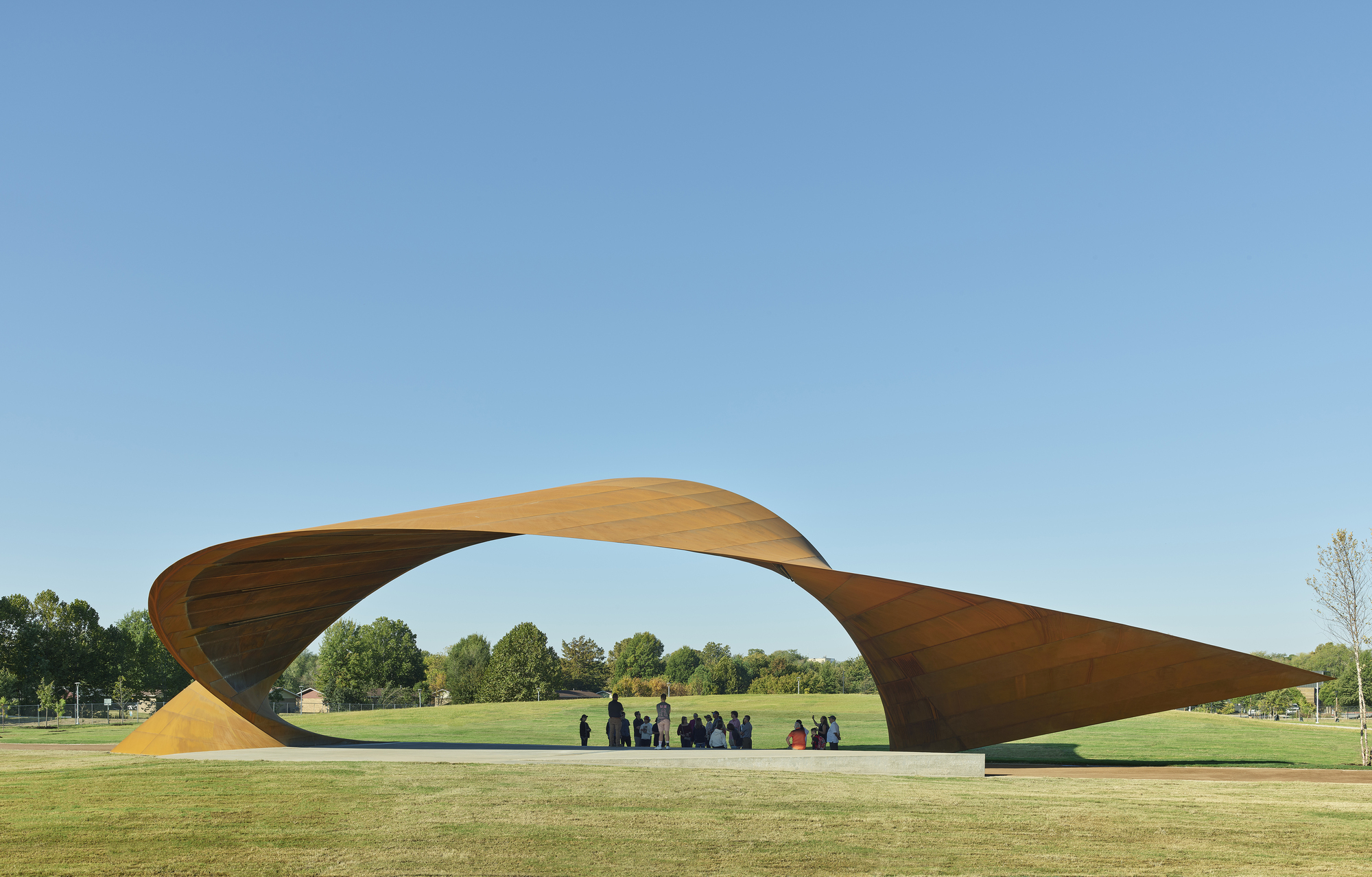 Twisted timber canopy spanning a lawn with visitors gathered beneath under clear blue sky
