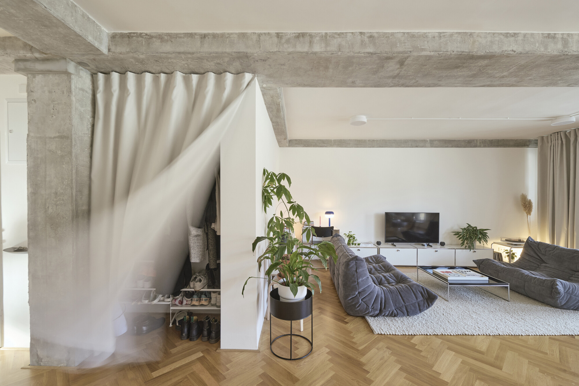 Living room with curtain partition concealing bedroom alcove beneath raw concrete structural elements