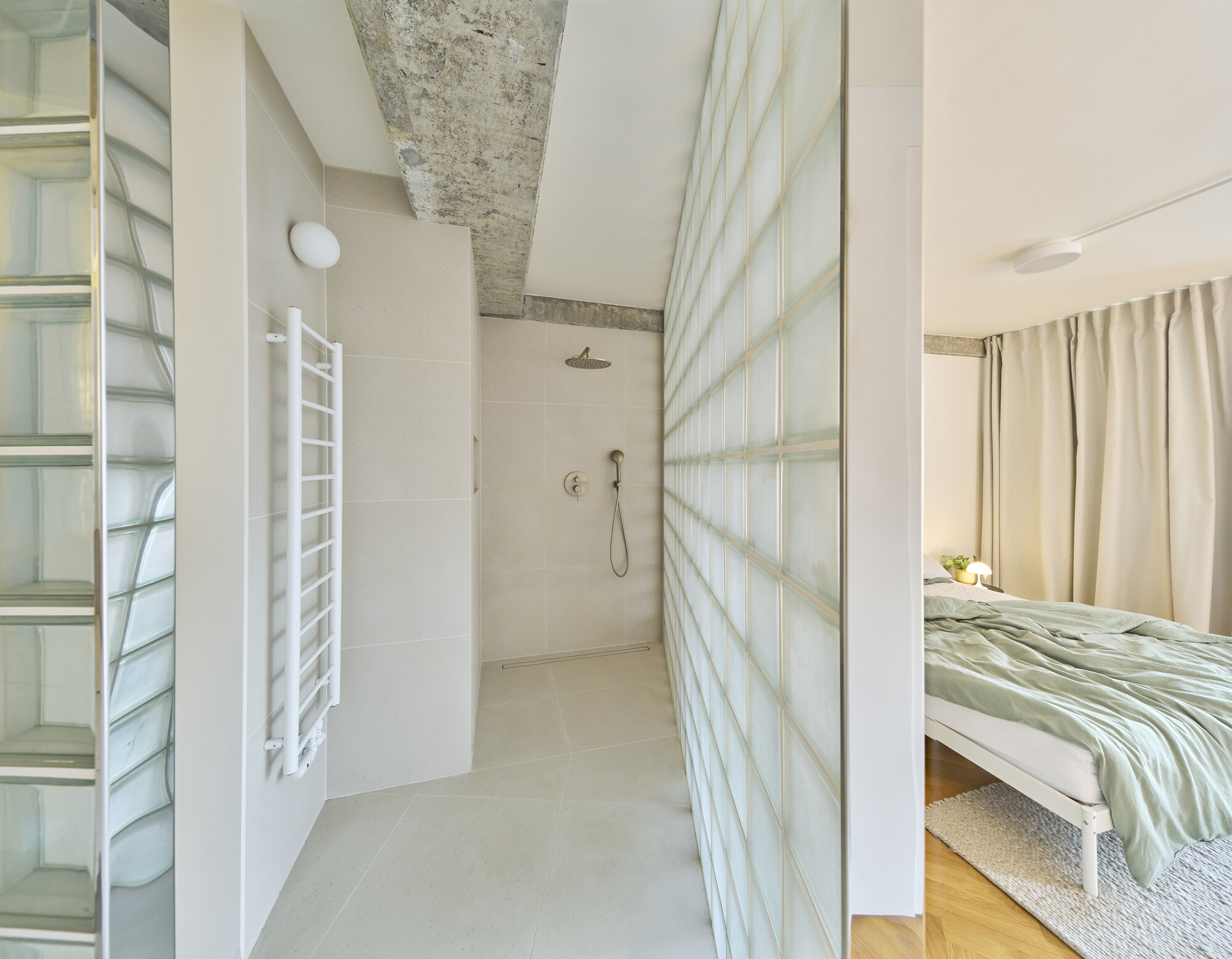 Bathroom with glass block wall and exposed concrete beam connecting to adjacent bedroom with curtain