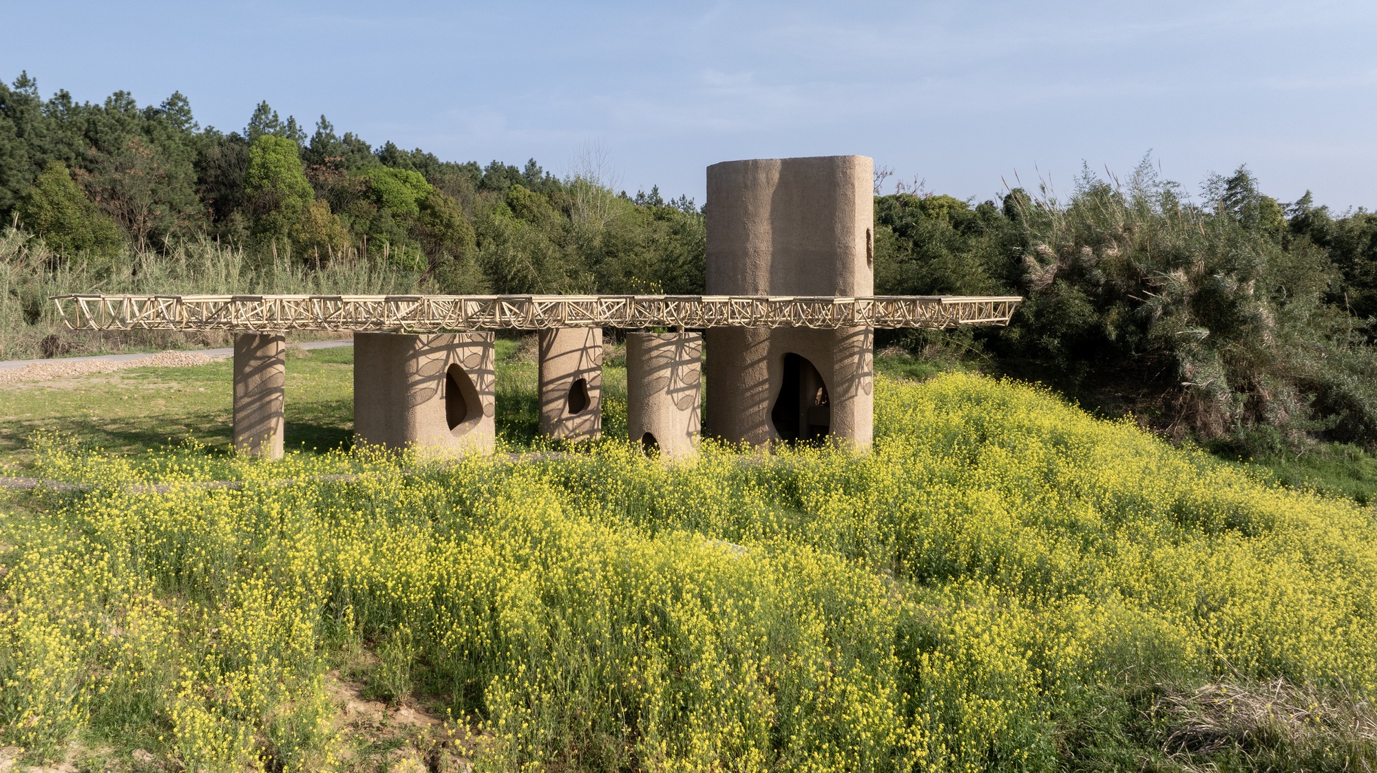 Cylindrical earthen columns with arched openings beneath a latticed canopy surrounded by blooming yellow wildflowers