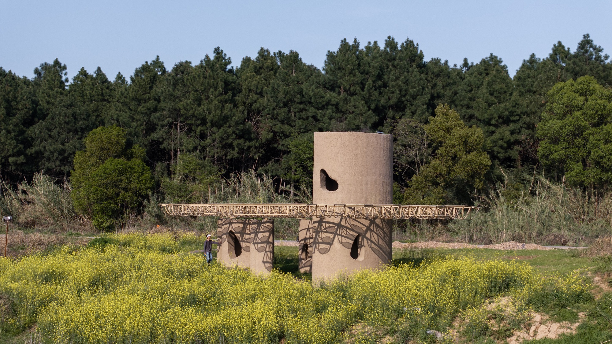 Cylindrical concrete tower with opening and timber canopy structure set among yellow wildflowers and dense forest backdrop
