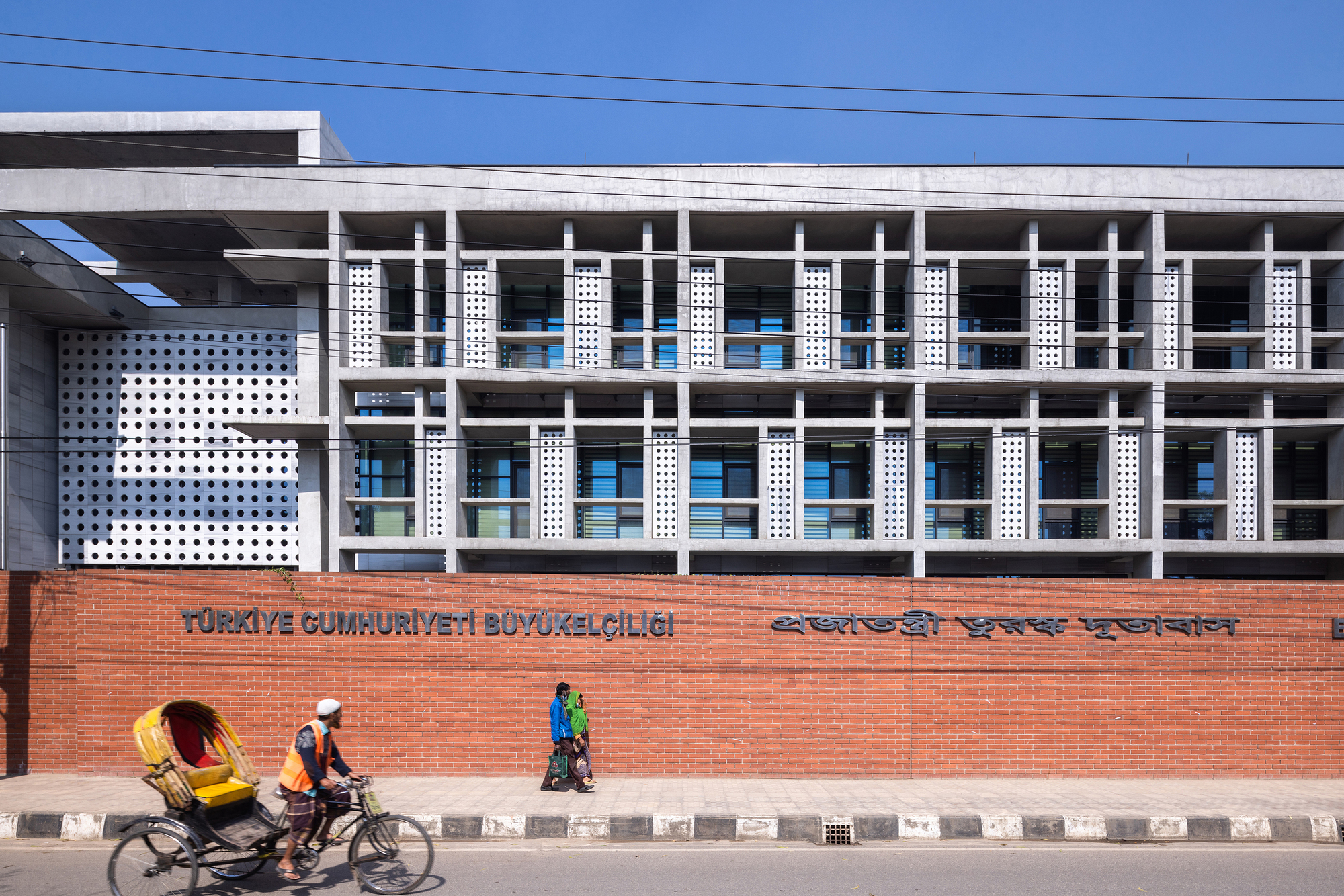 Street view of the perforated white facade with horizontal concrete frame above red brick boundary wall