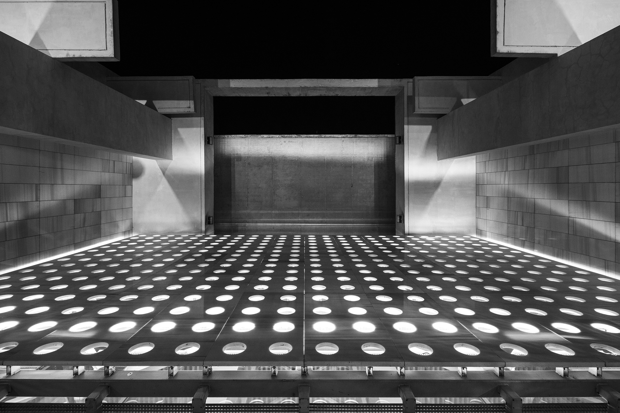 Upward view of perforated metal floor grate with circular openings and backlit concrete wall panel