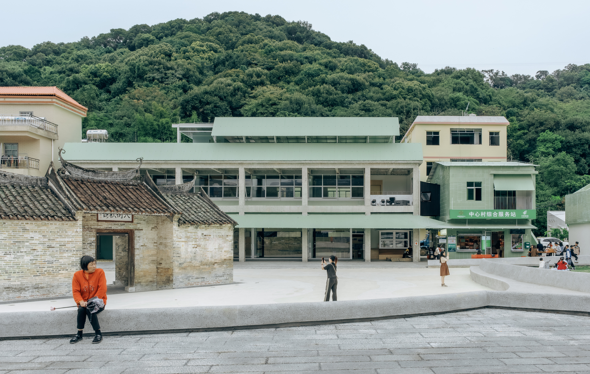Paved plaza with a traditional tile-roofed building beside a pale green multi-storey structure backed by forested hills