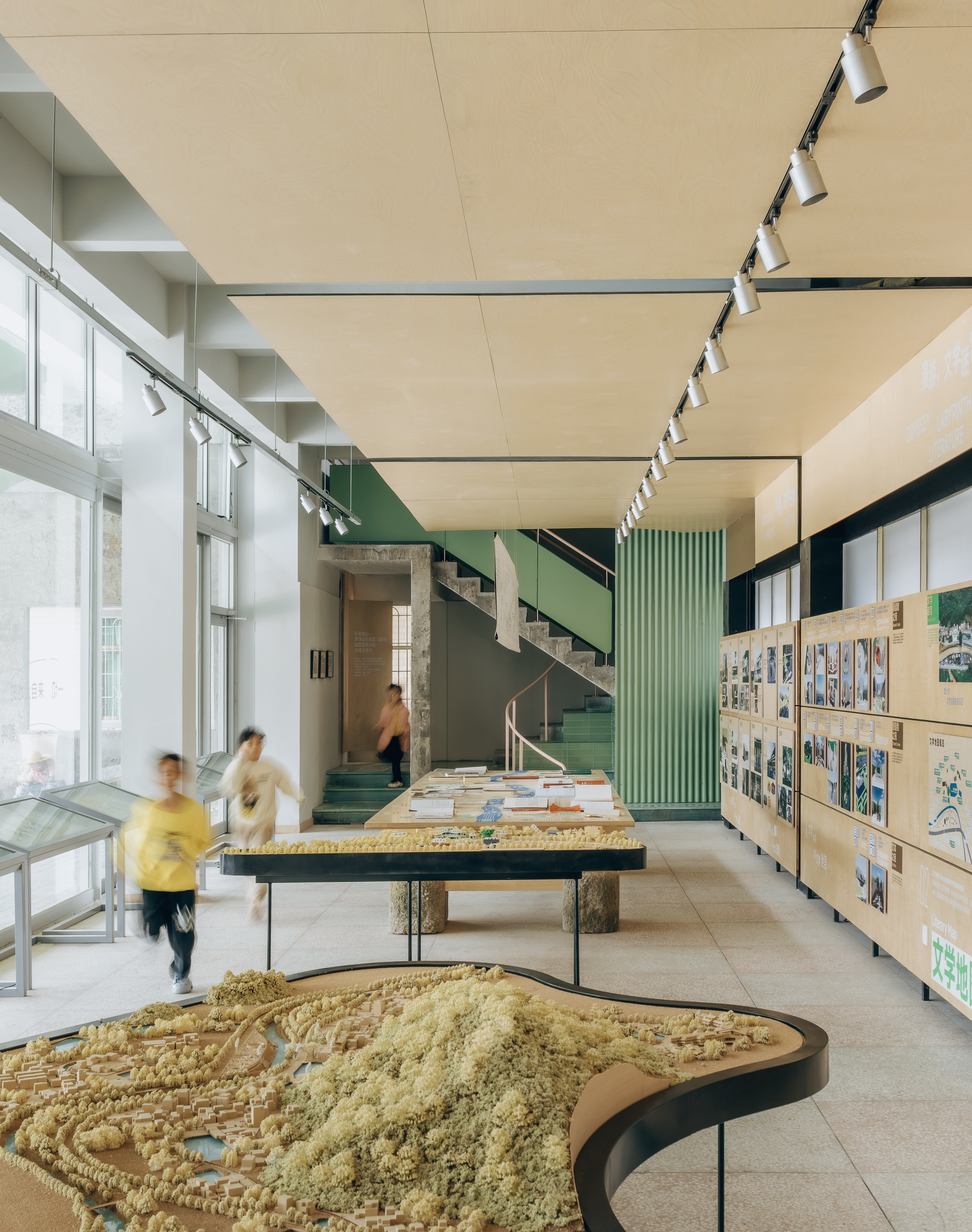 Exhibition hall interior with topographic models on tables, plywood display walls, and a green-sided staircase beyond