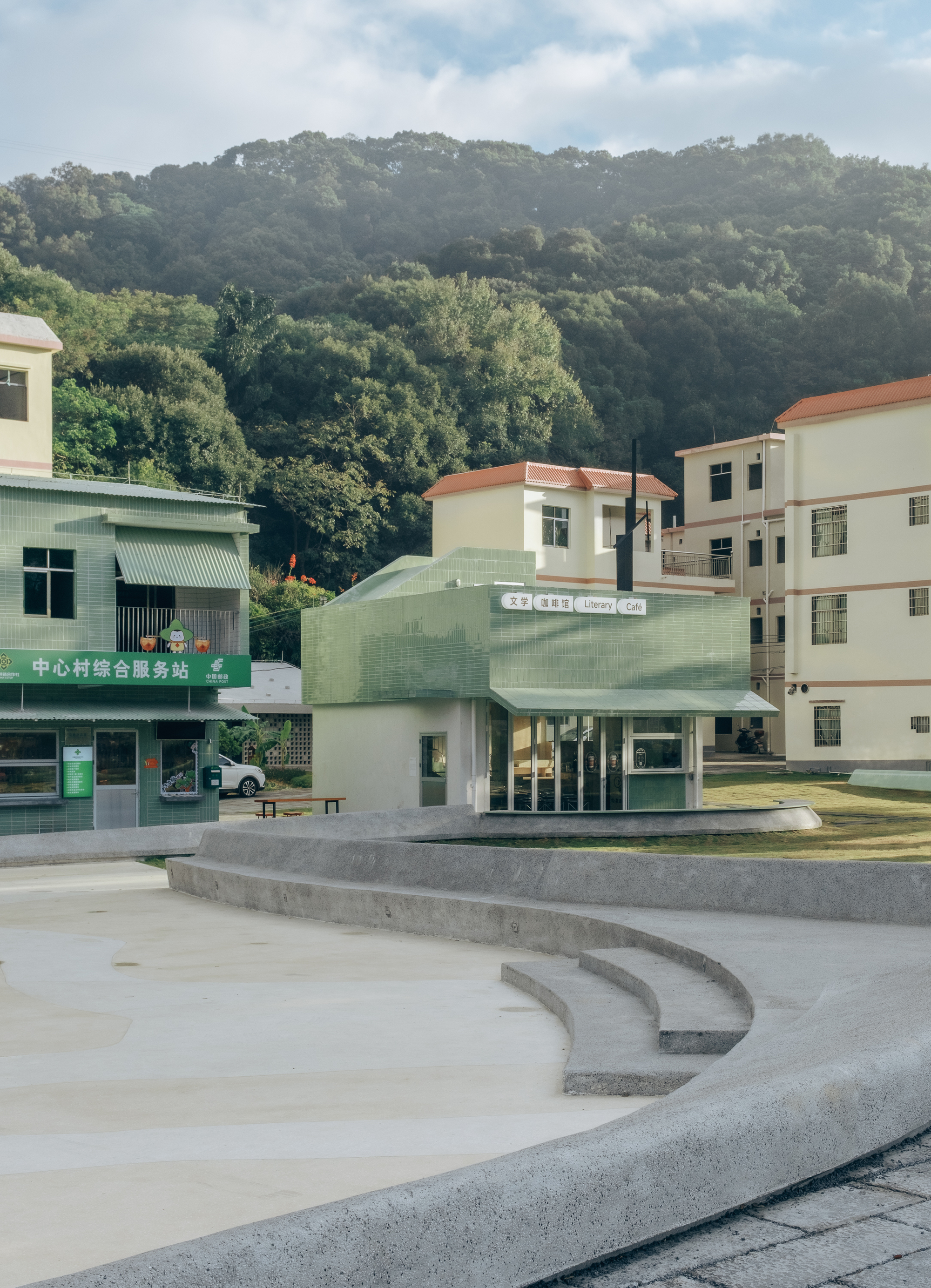 Curving concrete terraces in a plaza facing a compact pale green library pavilion with tile-clad upper volume