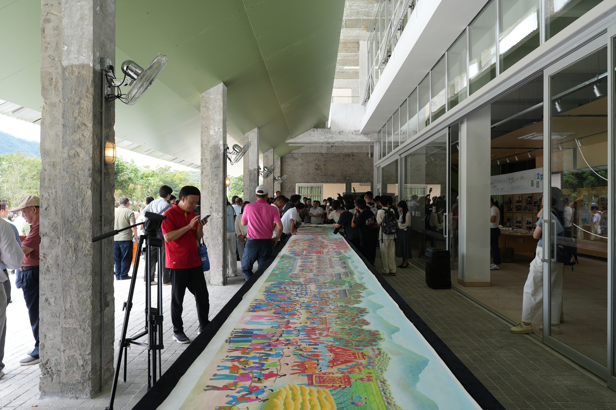 Covered exterior corridor with concrete columns and visitors viewing a long painted mural on the floor