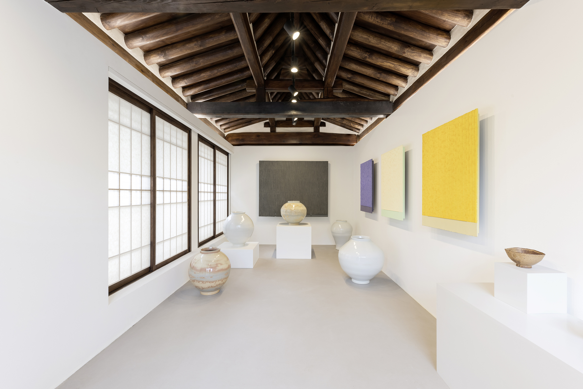 Gallery room with white ceramic vessels on pedestals and colorful paintings beneath timber truss ceiling