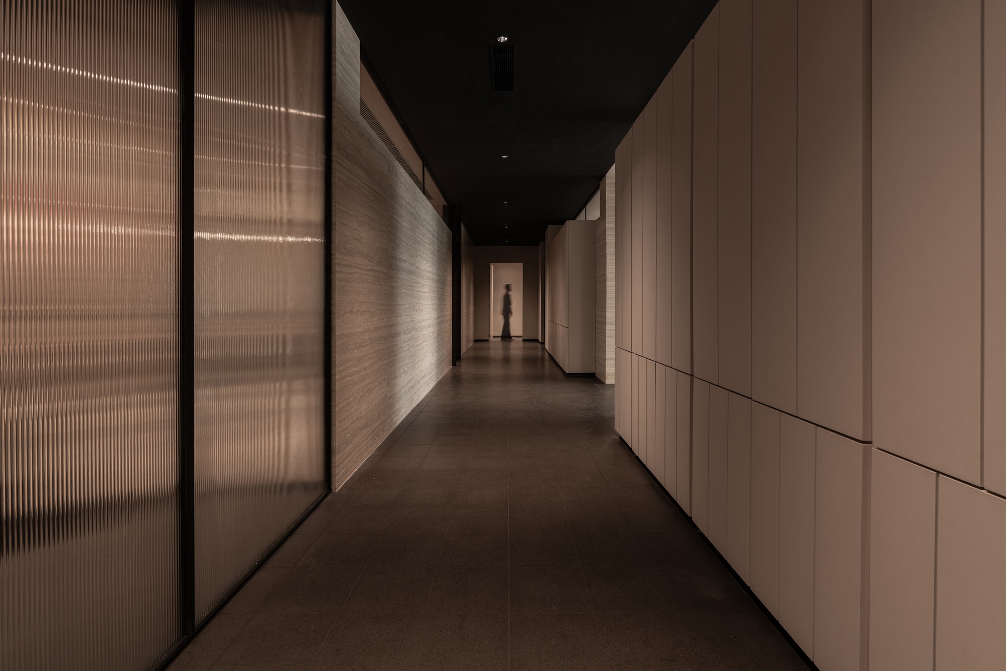 Narrow hallway with ribbed translucent glass walls and wood paneling leading to a distant silhouette