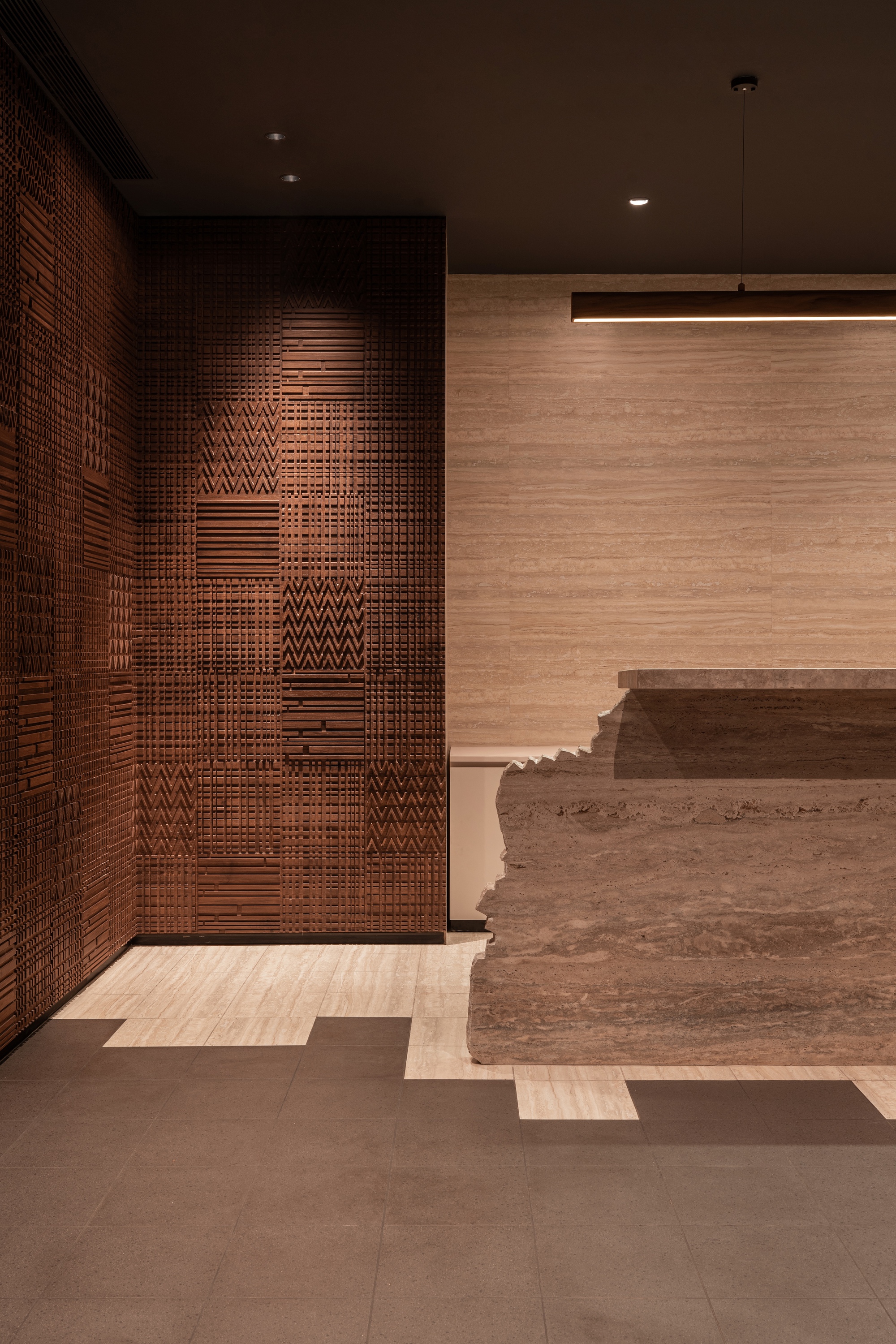 Textured copper tile wall adjacent to travertine reception desk with horizontal pendant light fixture