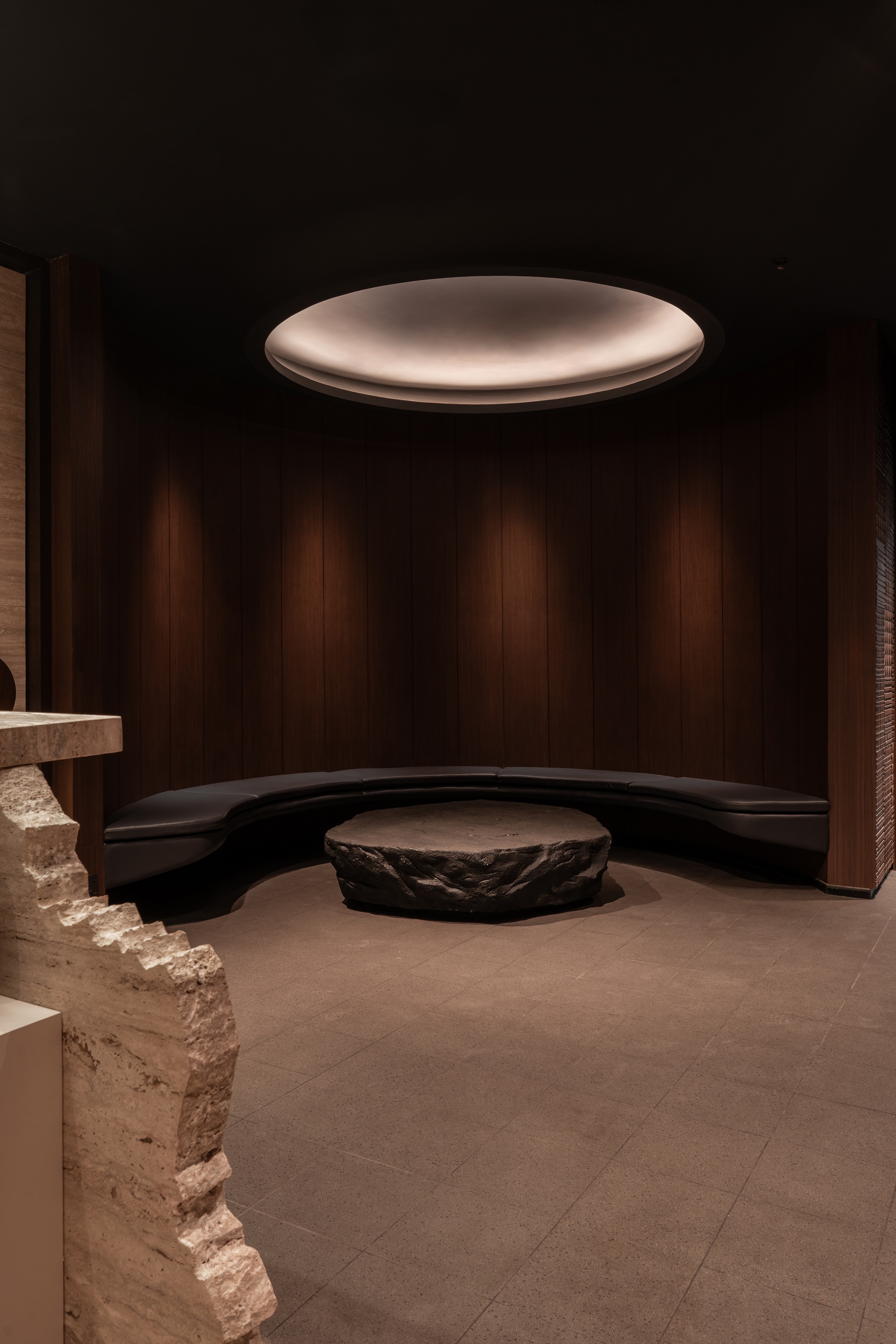 Curved black bench and raw stone table beneath glowing disc ceiling fixture in dark room