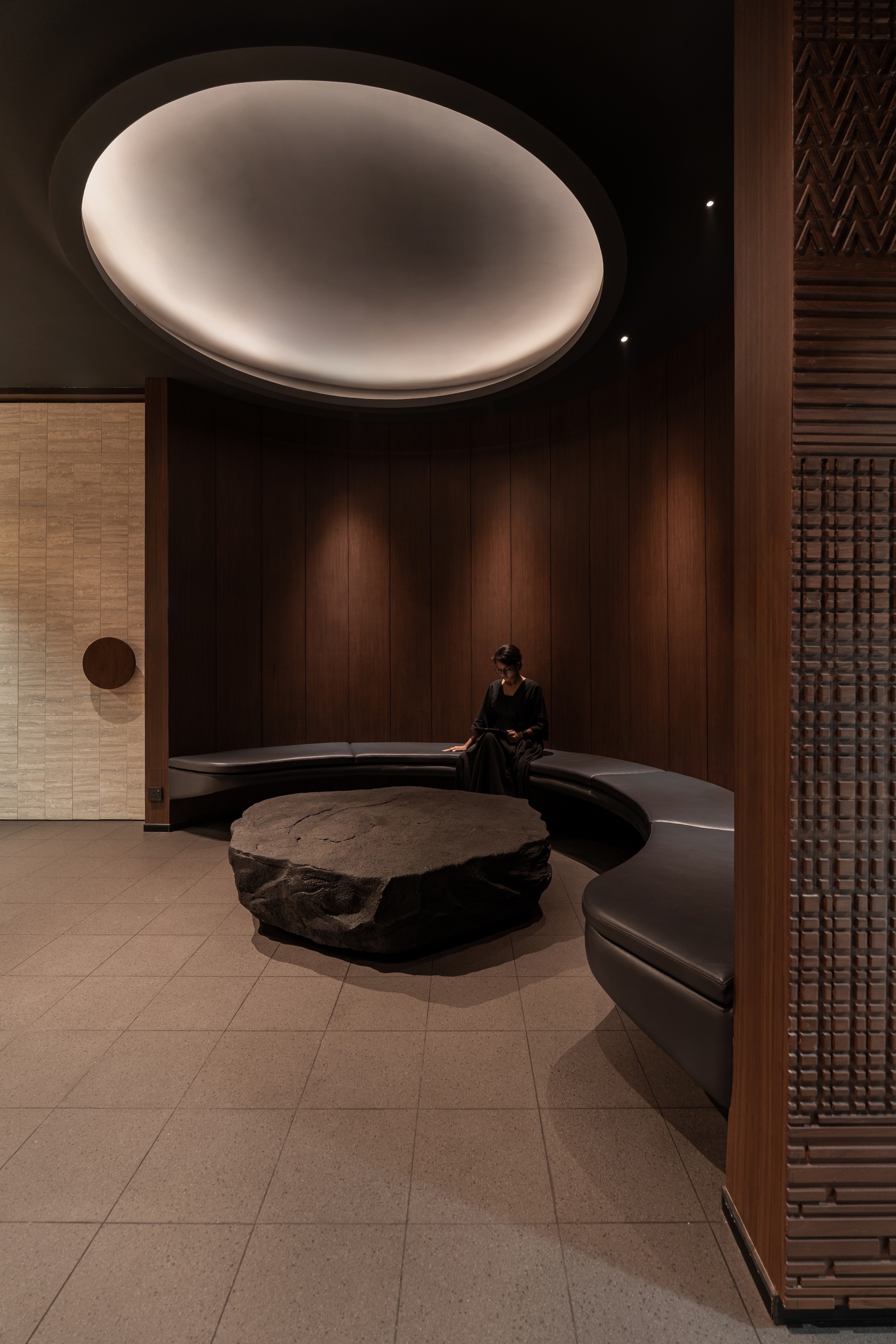 Person seated on curved bench below illuminated disc ceiling element and textured timber screens