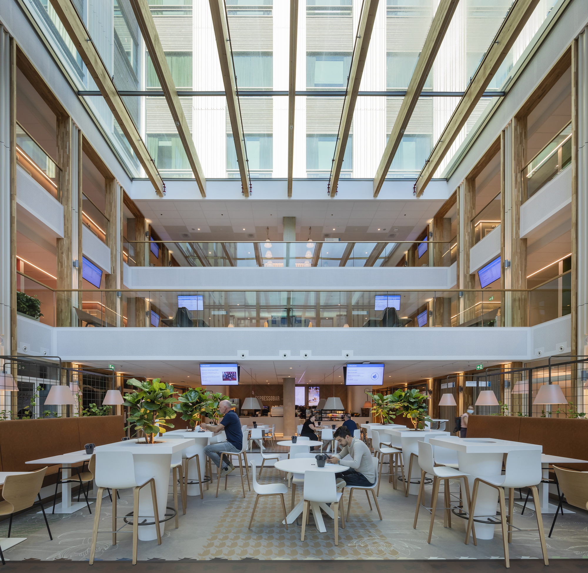 Multi-level atrium cafe with angled glass roof and people seated at white tables