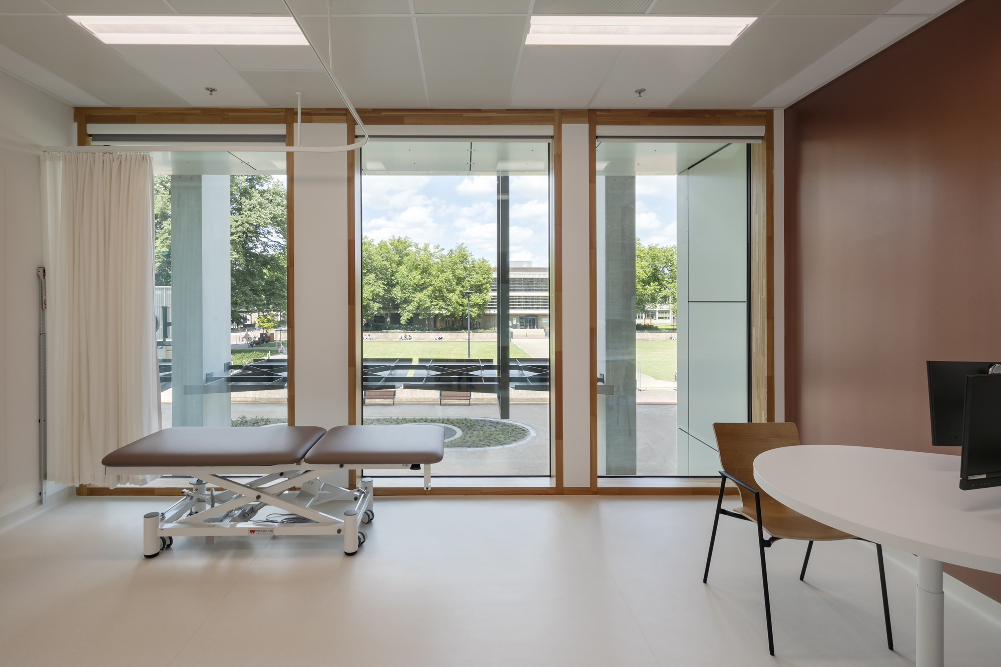 Treatment room with timber-framed windows overlooking a tree-lined plaza in summer