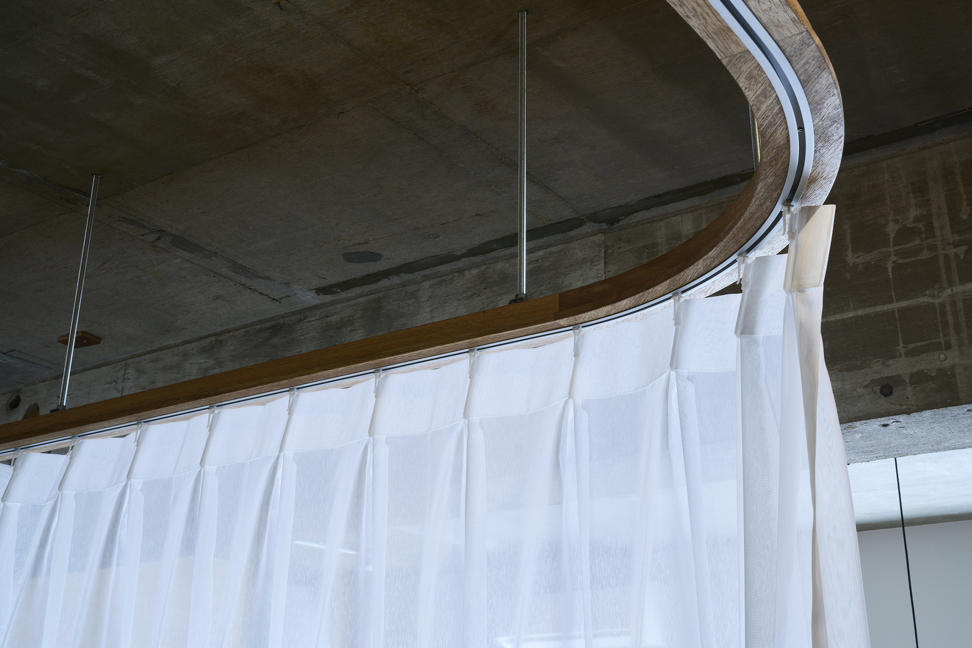 Close-up of curved timber rail with white curtain suspended from exposed concrete ceiling