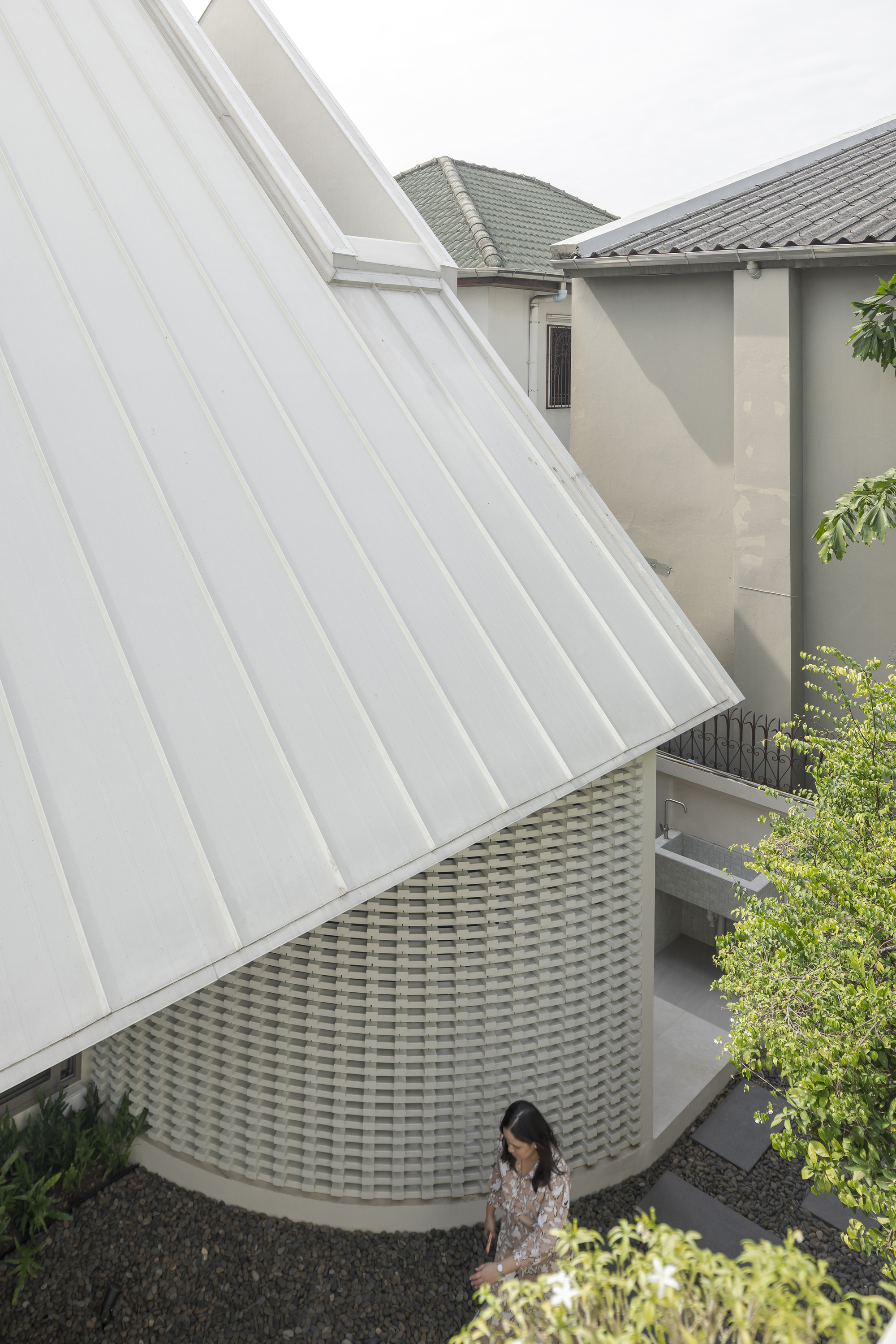 White ribbed metal roof plane rising above a woven brick wall with a woman walking below