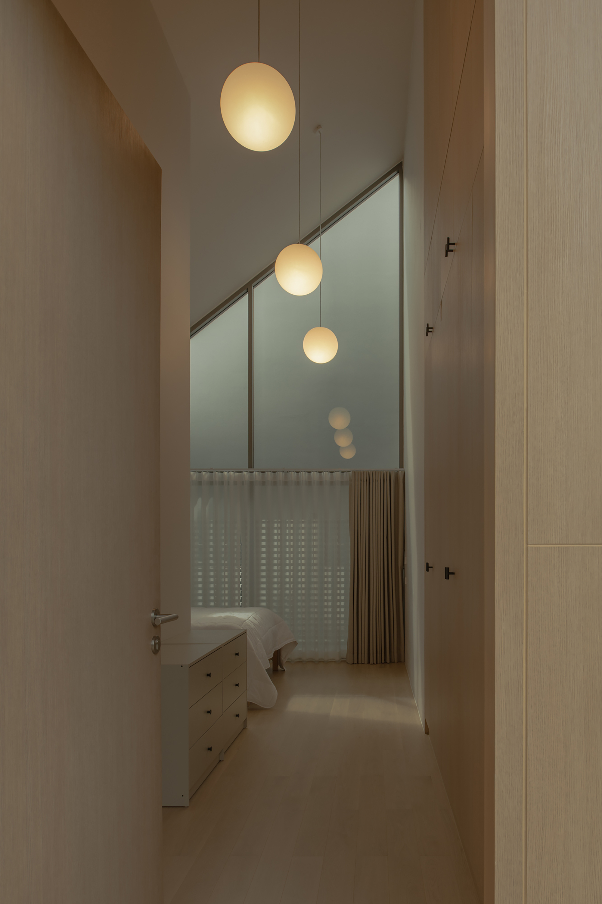 Corridor view through angled glass partition with suspended globe pendants and translucent curtain