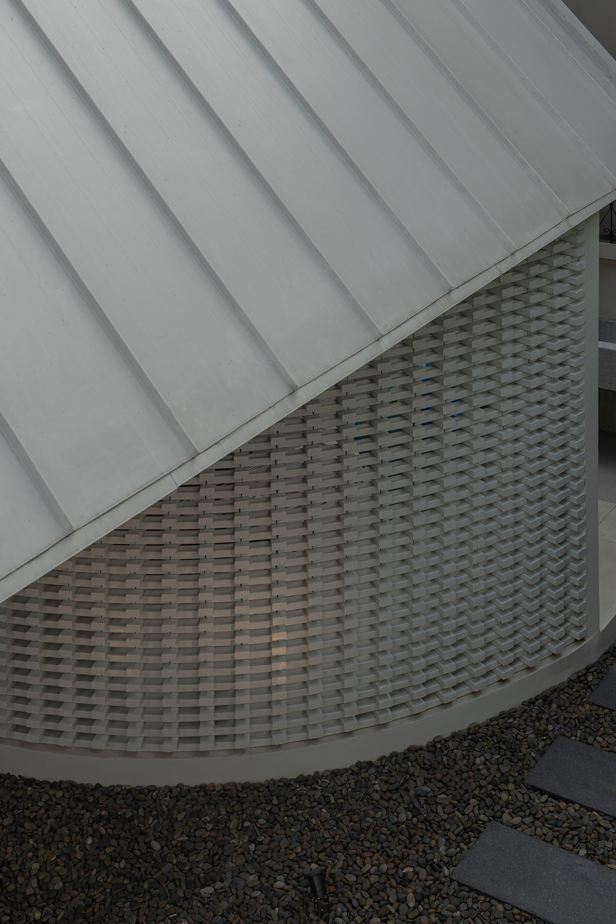 Detail of curved woven brick facade beneath metal standing seam roof and gravel courtyard