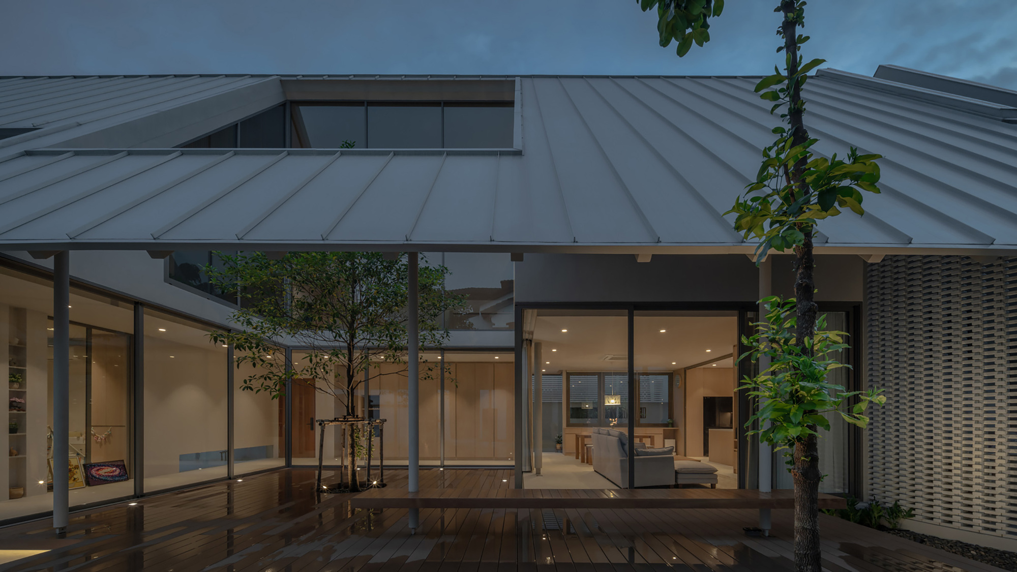 Courtyard terrace with timber deck and planted trees enclosed by glass walls beneath a standing seam metal roof