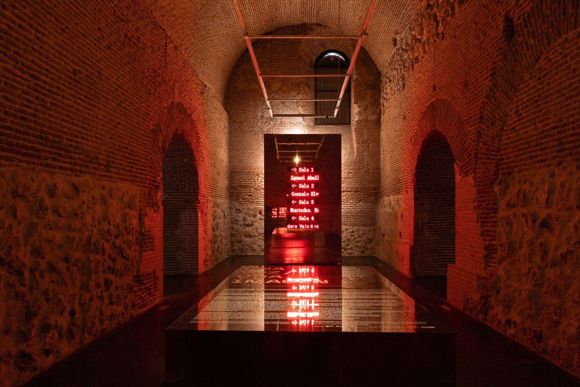 Vaulted brick corridor with red LED signage reflected in a water channel running down the center