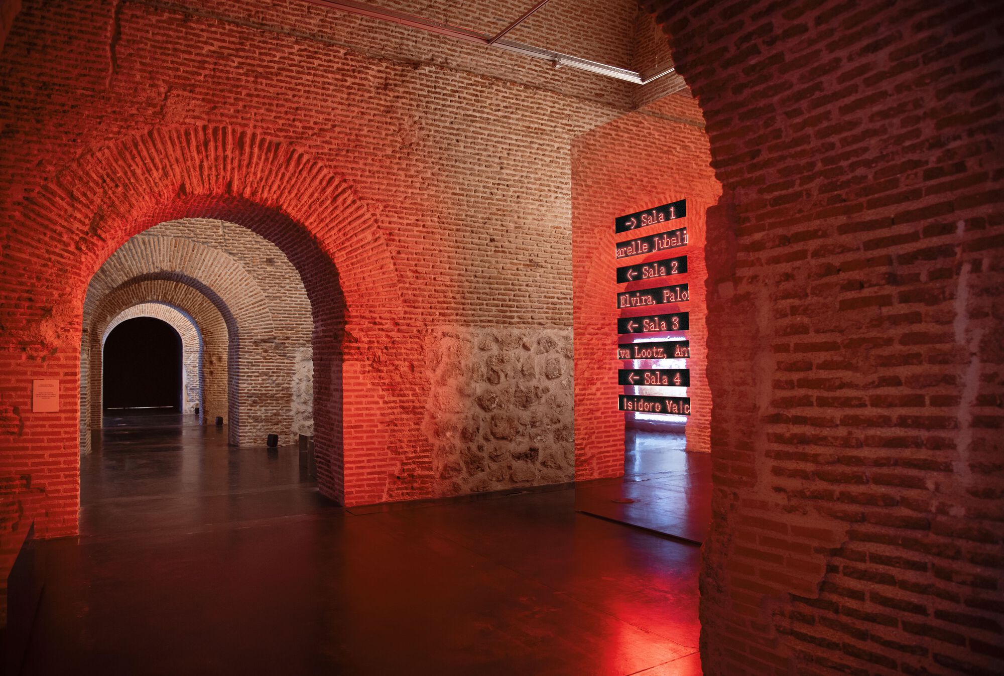 Sequence of brick arches receding through red-lit chambers with stone and brick walls