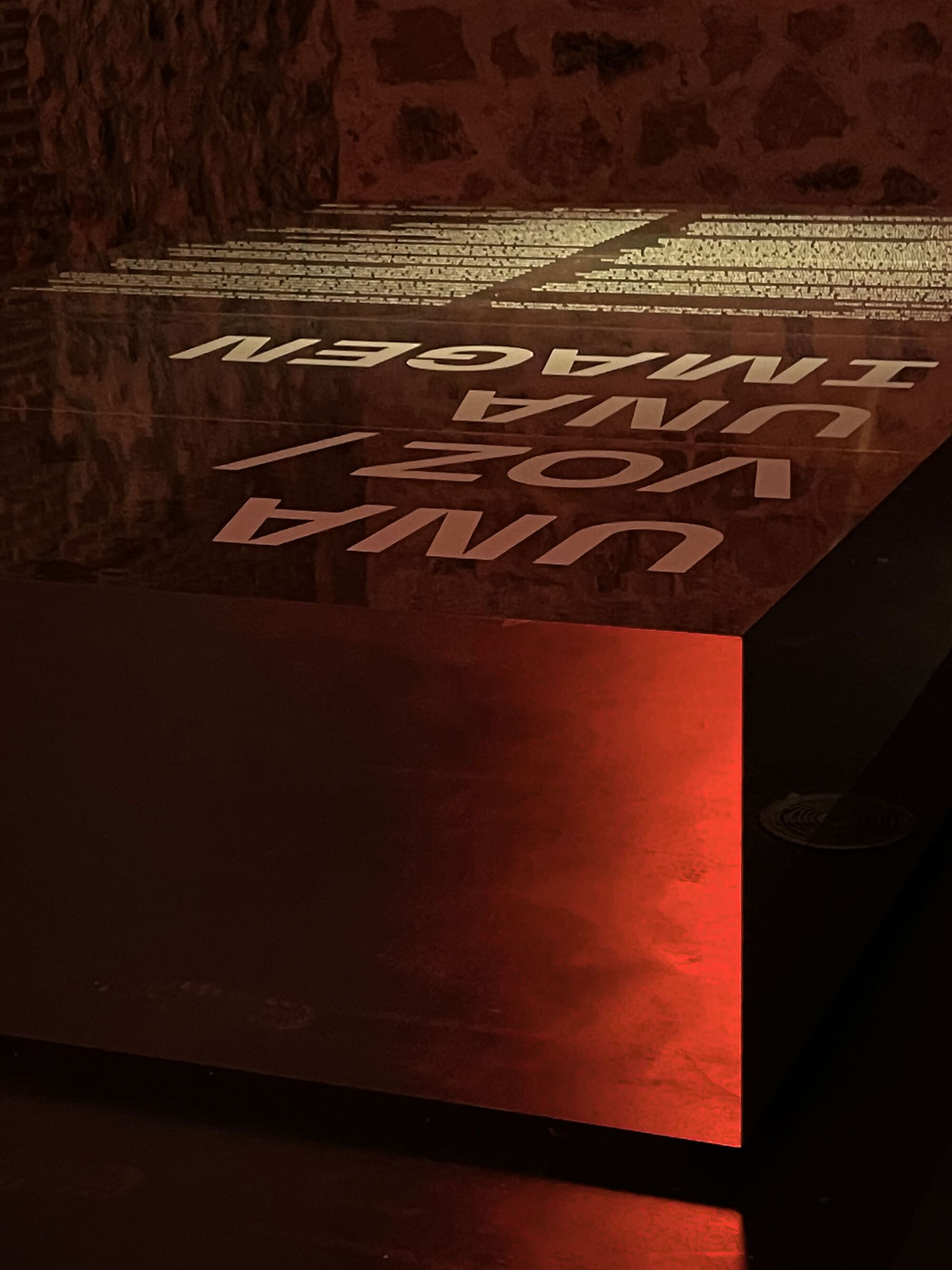 Angled view of an illuminated red metal panel with reversed text against a textured wall