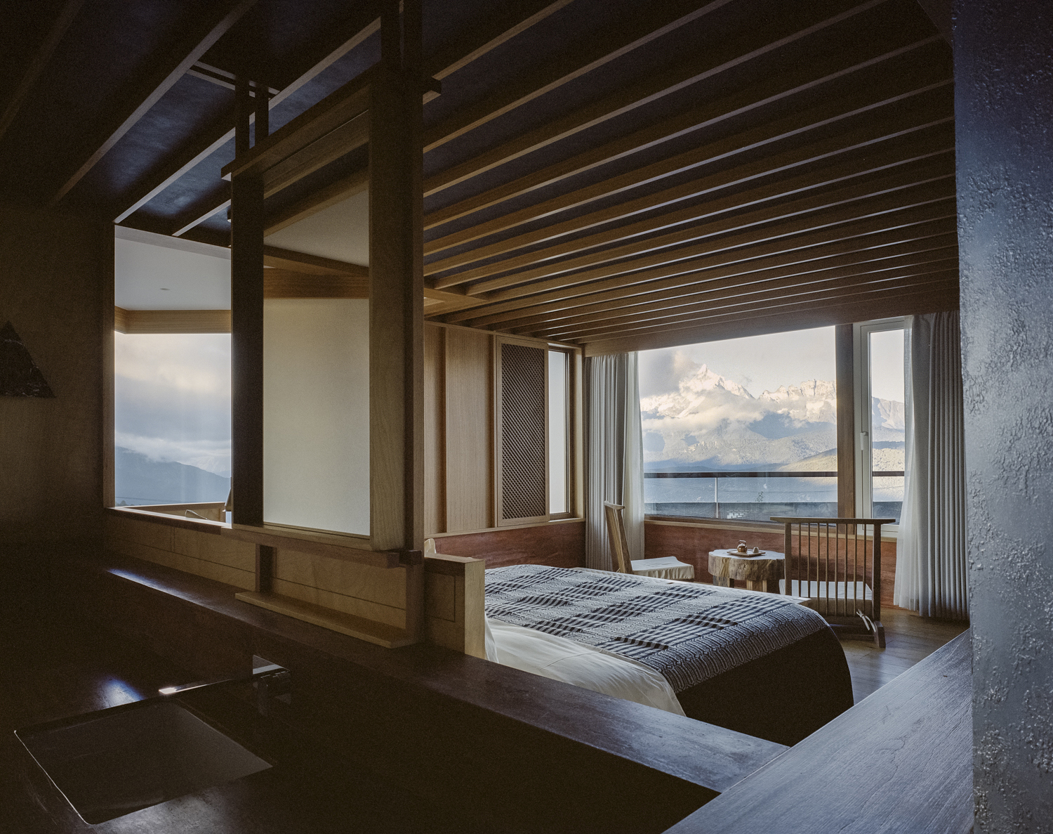 Bedroom with exposed timber beams and full-height windows facing distant clouds and lake