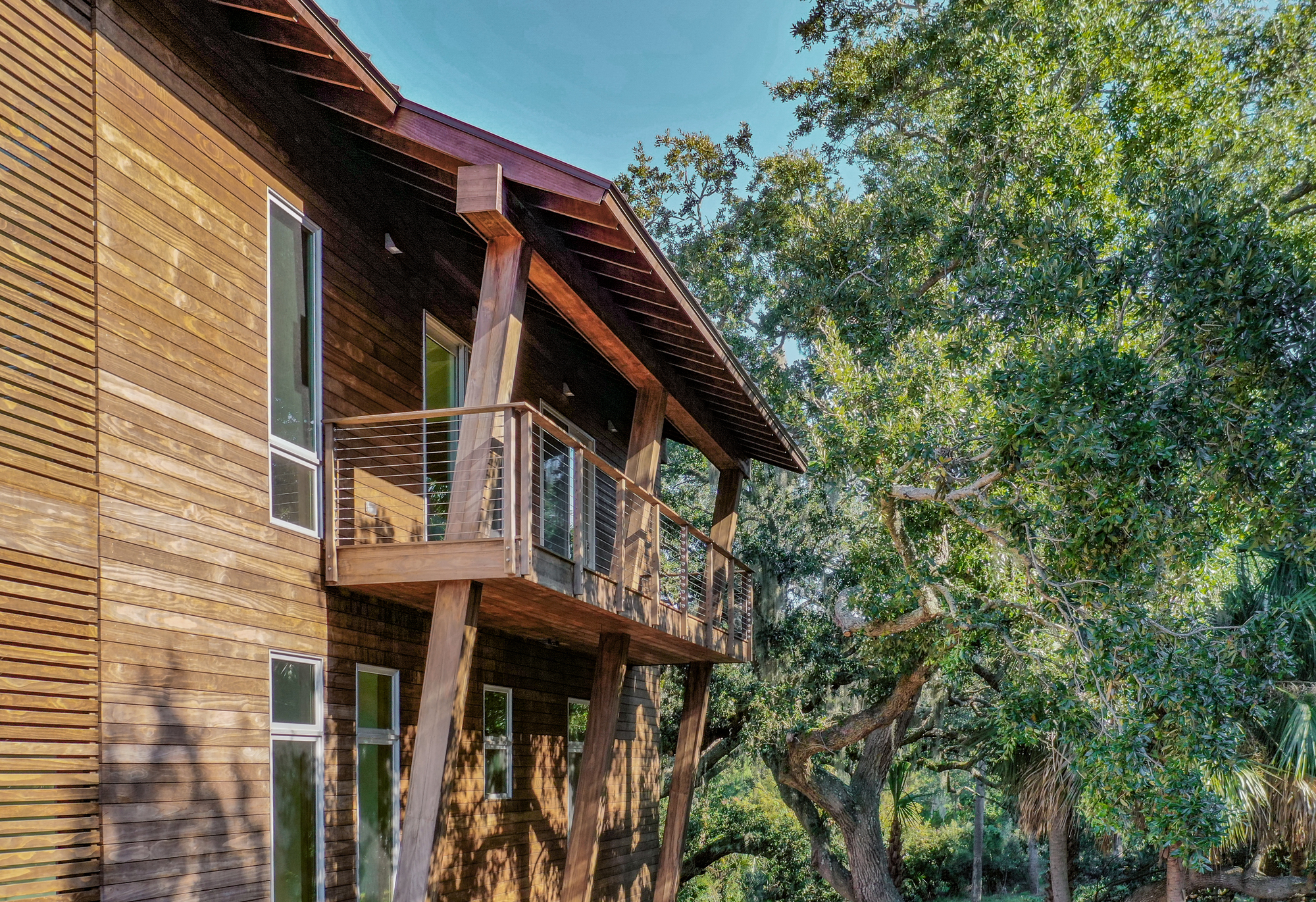 Two-story timber facade with horizontal siding and cantilevered balcony beneath mature oak trees