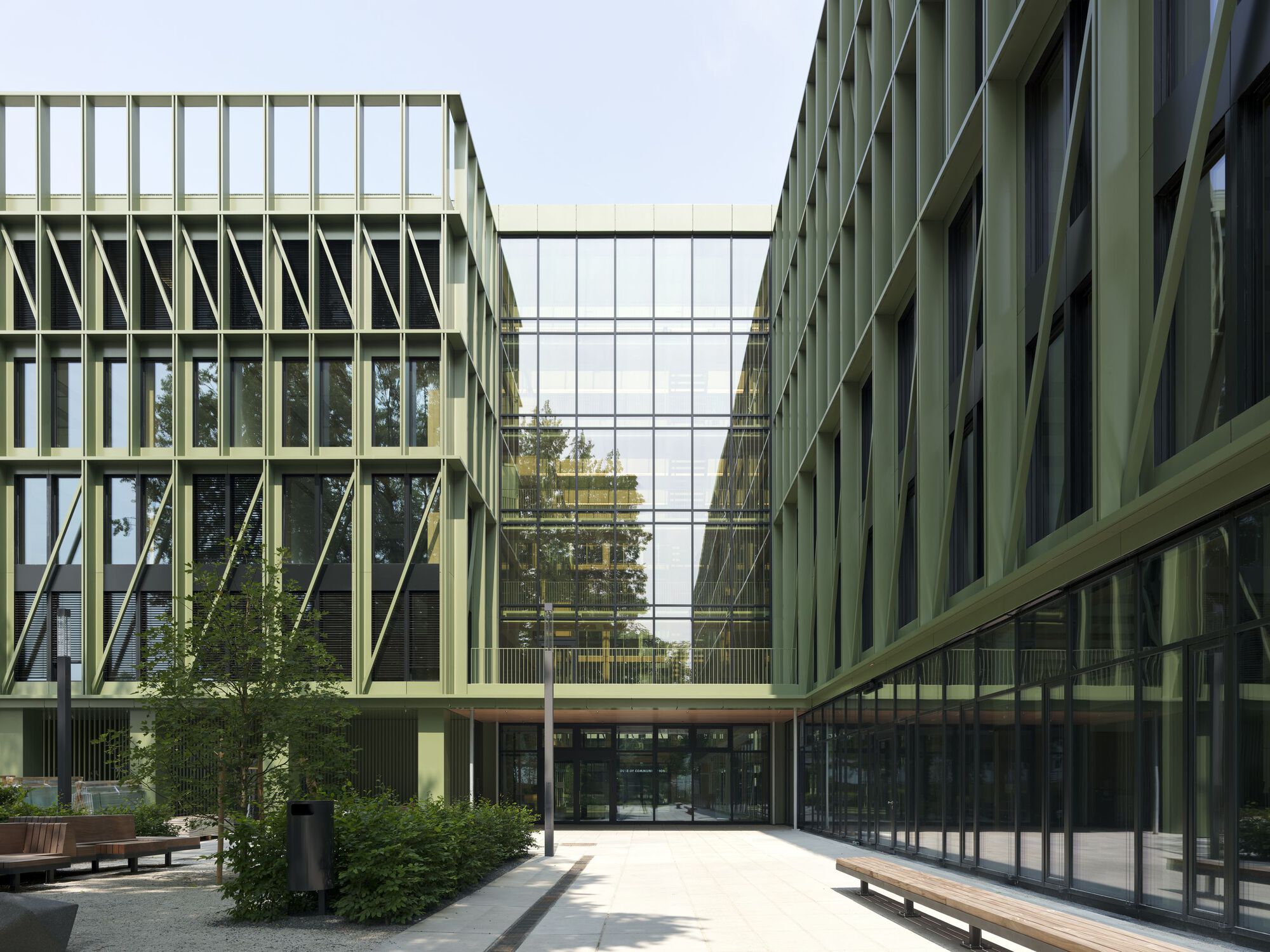 Glass skybridge connecting two wings with diagonal bracing over a courtyard entry and wooden benches