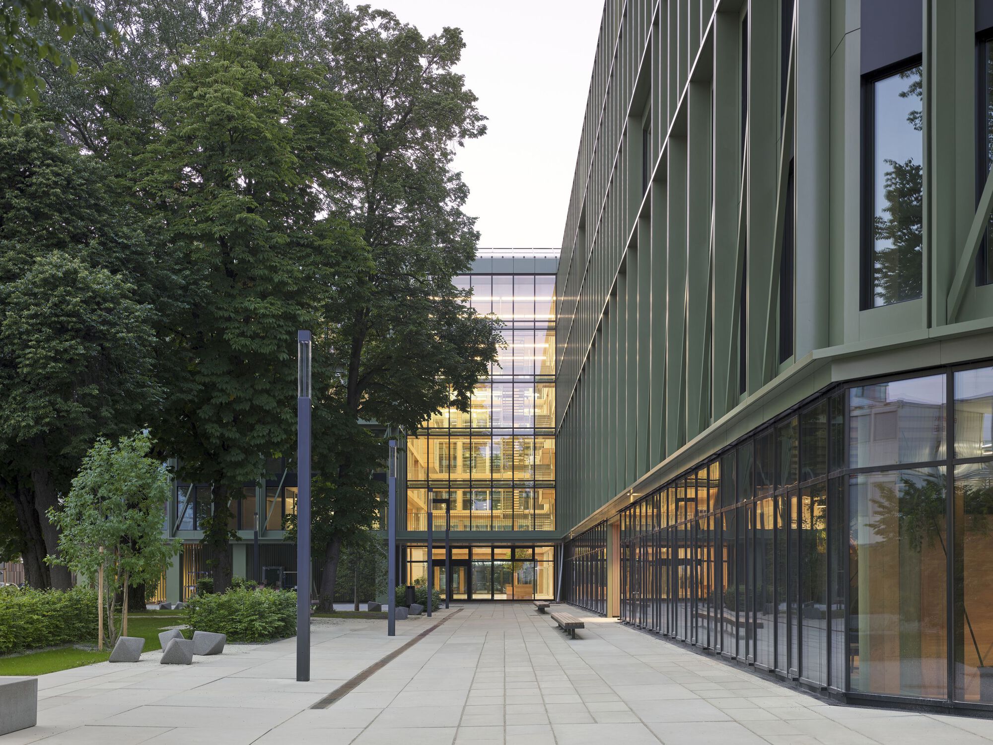 Paved plaza between two volumes with vertical green cladding and mature trees at dusk