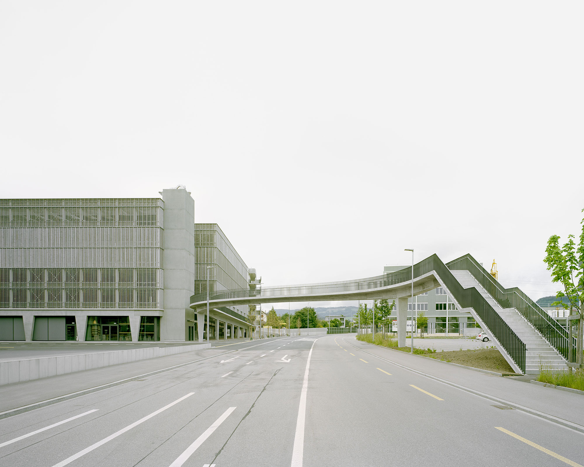 Pedestrian bridge crossing the roadway between two glass and concrete volumes under an overcast sky