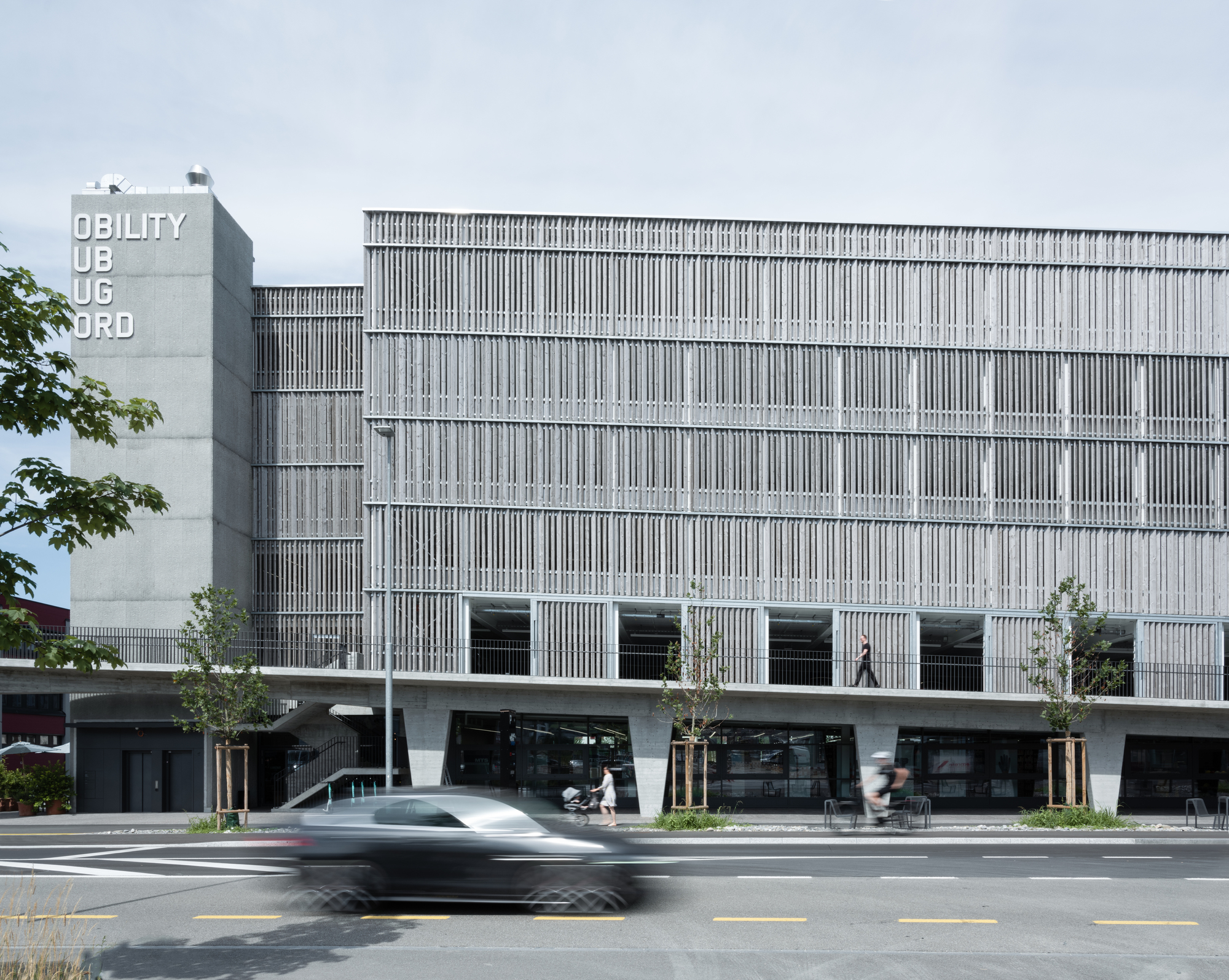 Facade with vertical louvers and canted concrete columns at street level as a car passes in the foreground