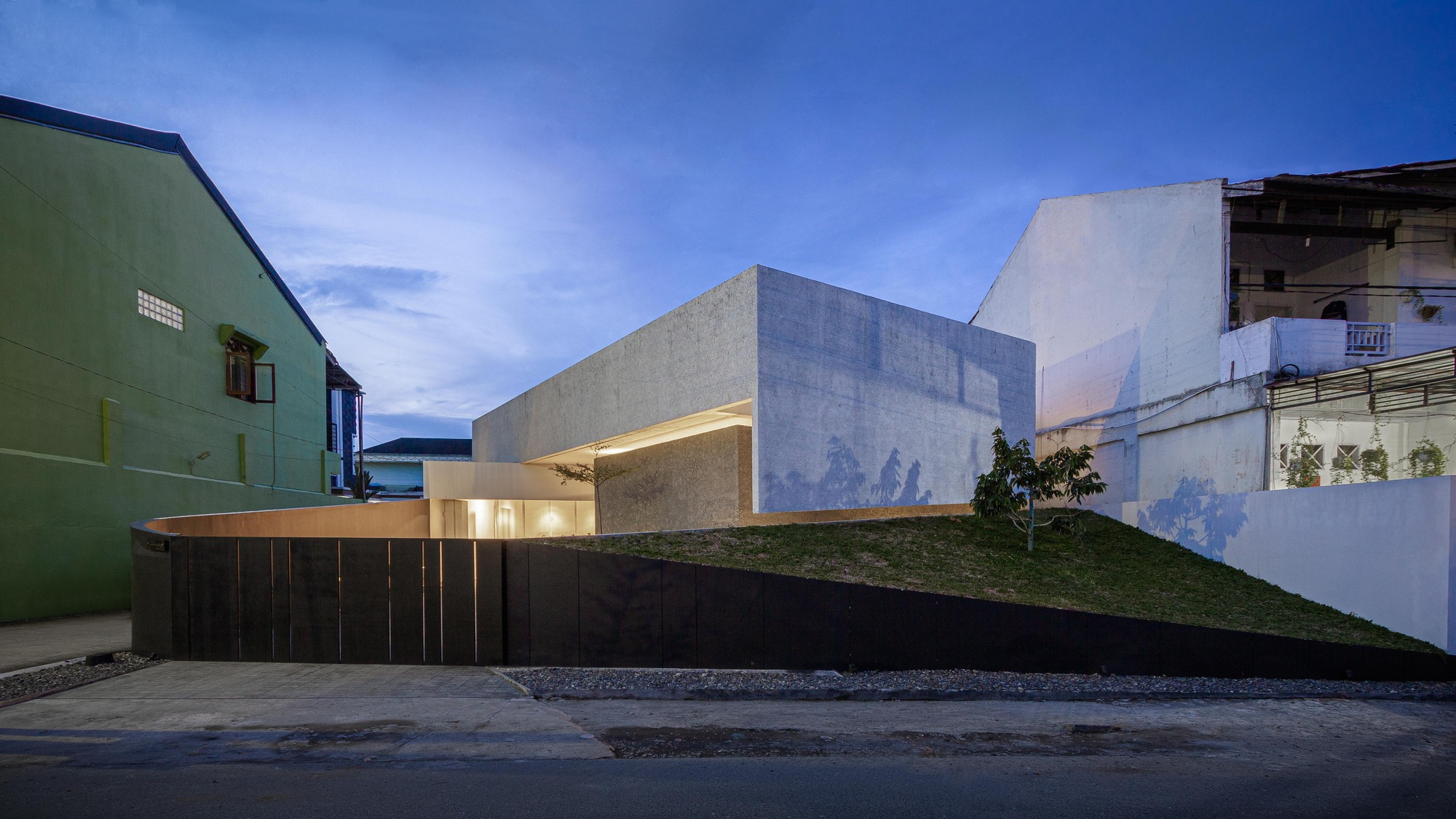 Angled concrete volumes with grass-covered ramp and black timber cladding against a blue evening sky