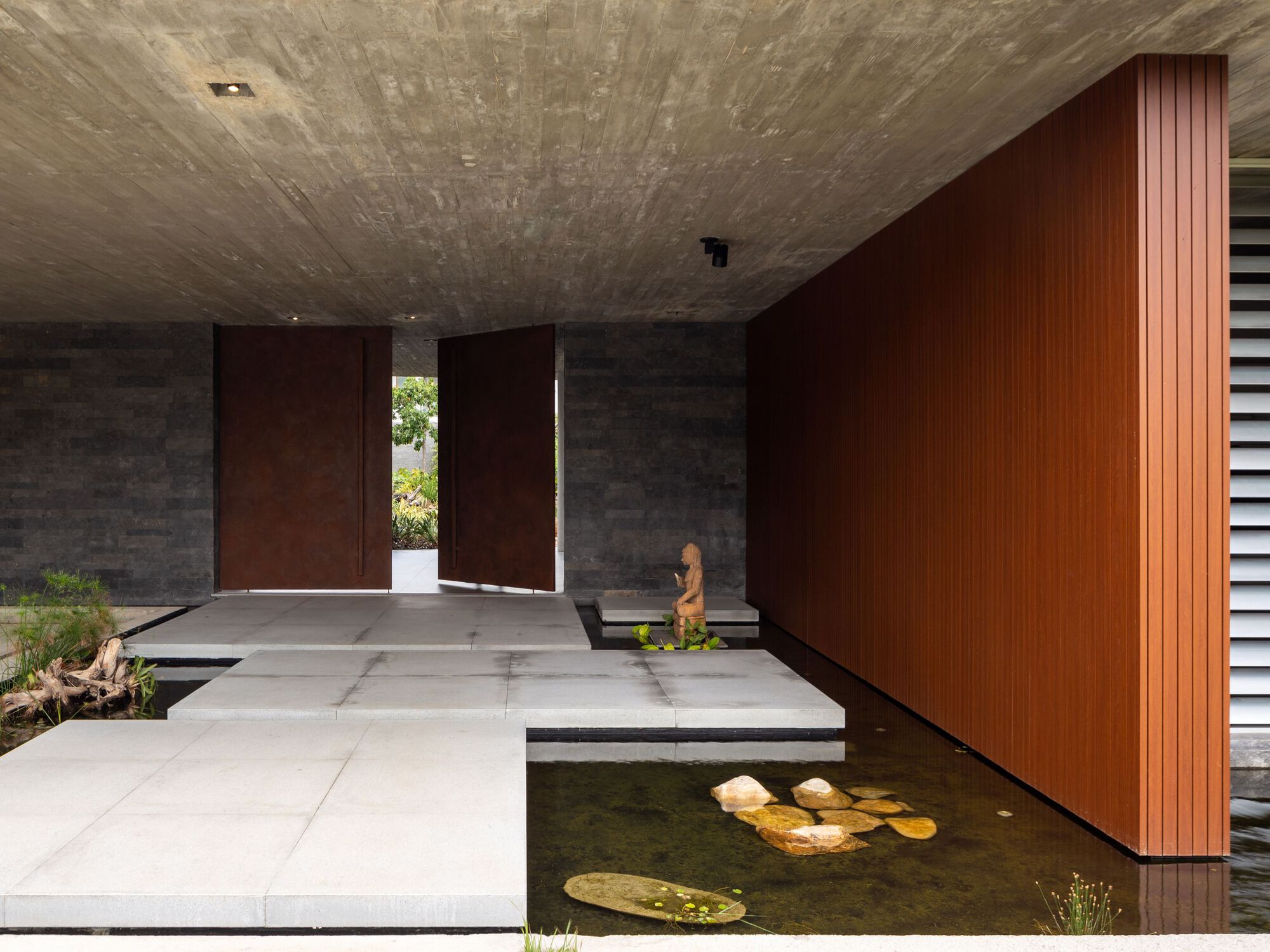 Covered entrance with board-formed concrete ceiling, timber-clad wall, and stepping stones over a reflecting pool