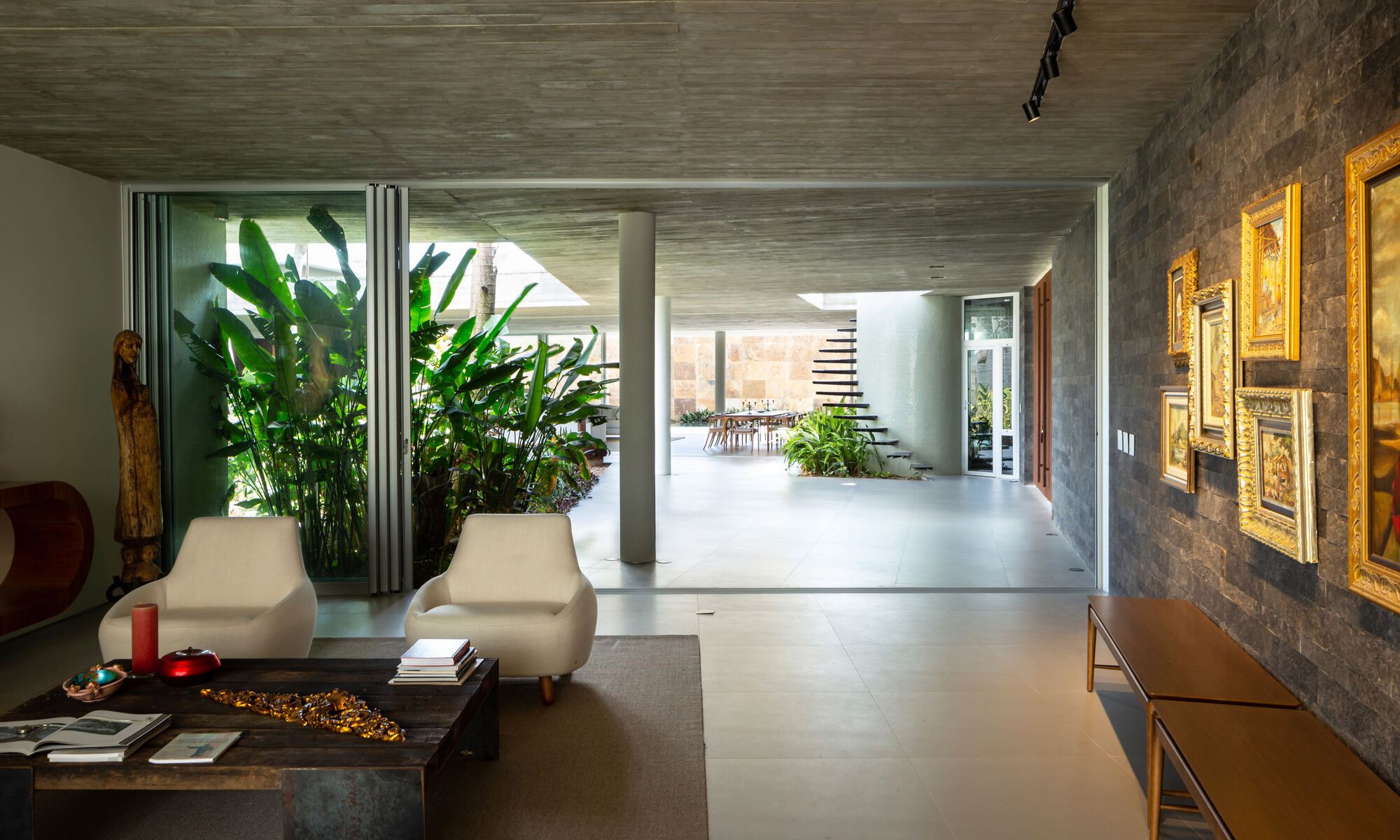 Living room view through to interior courtyard planted with tall banana palms under exposed concrete ceiling
