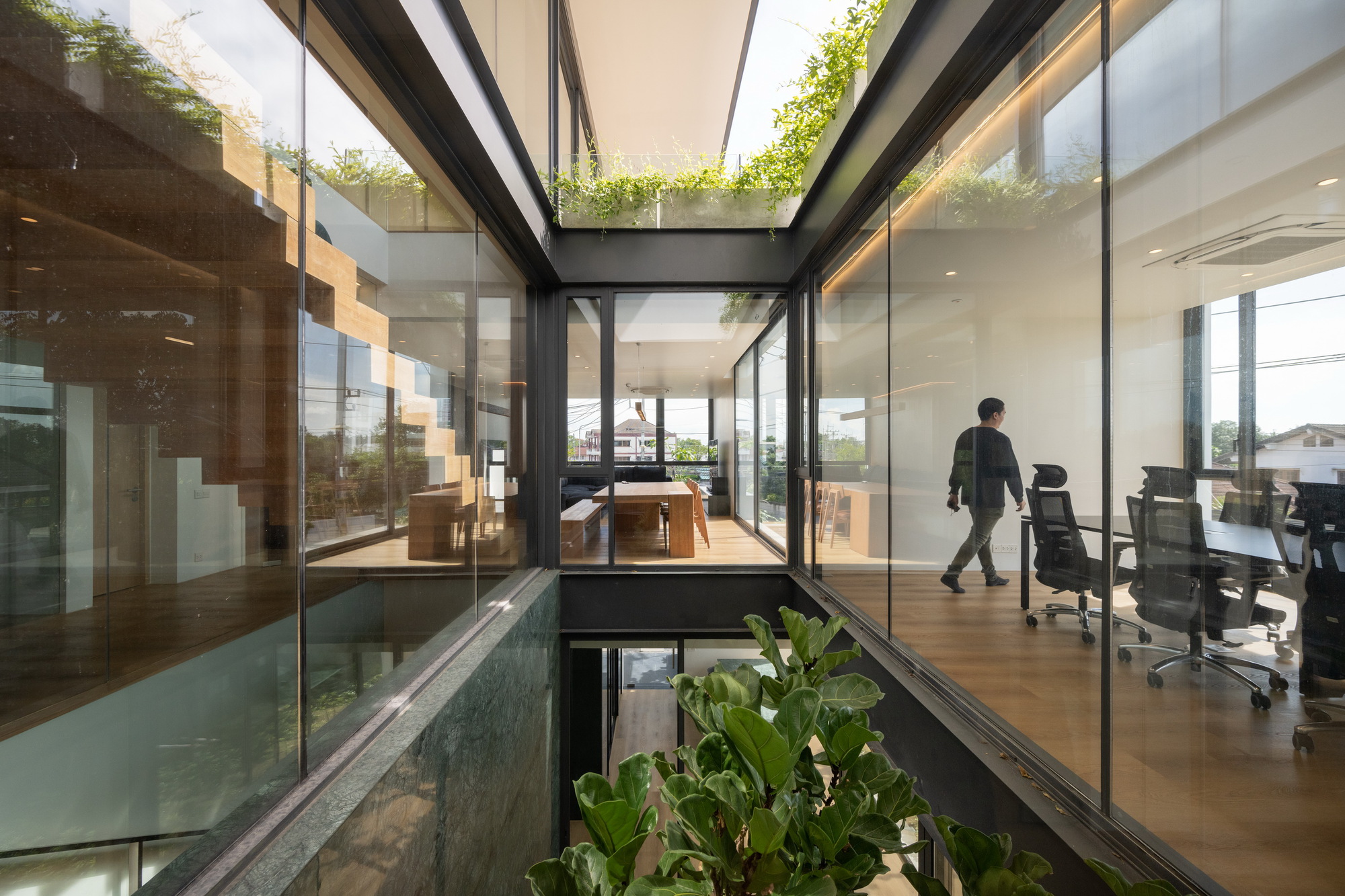 Glass-enclosed void with floating timber staircase visible from office space as person walks by